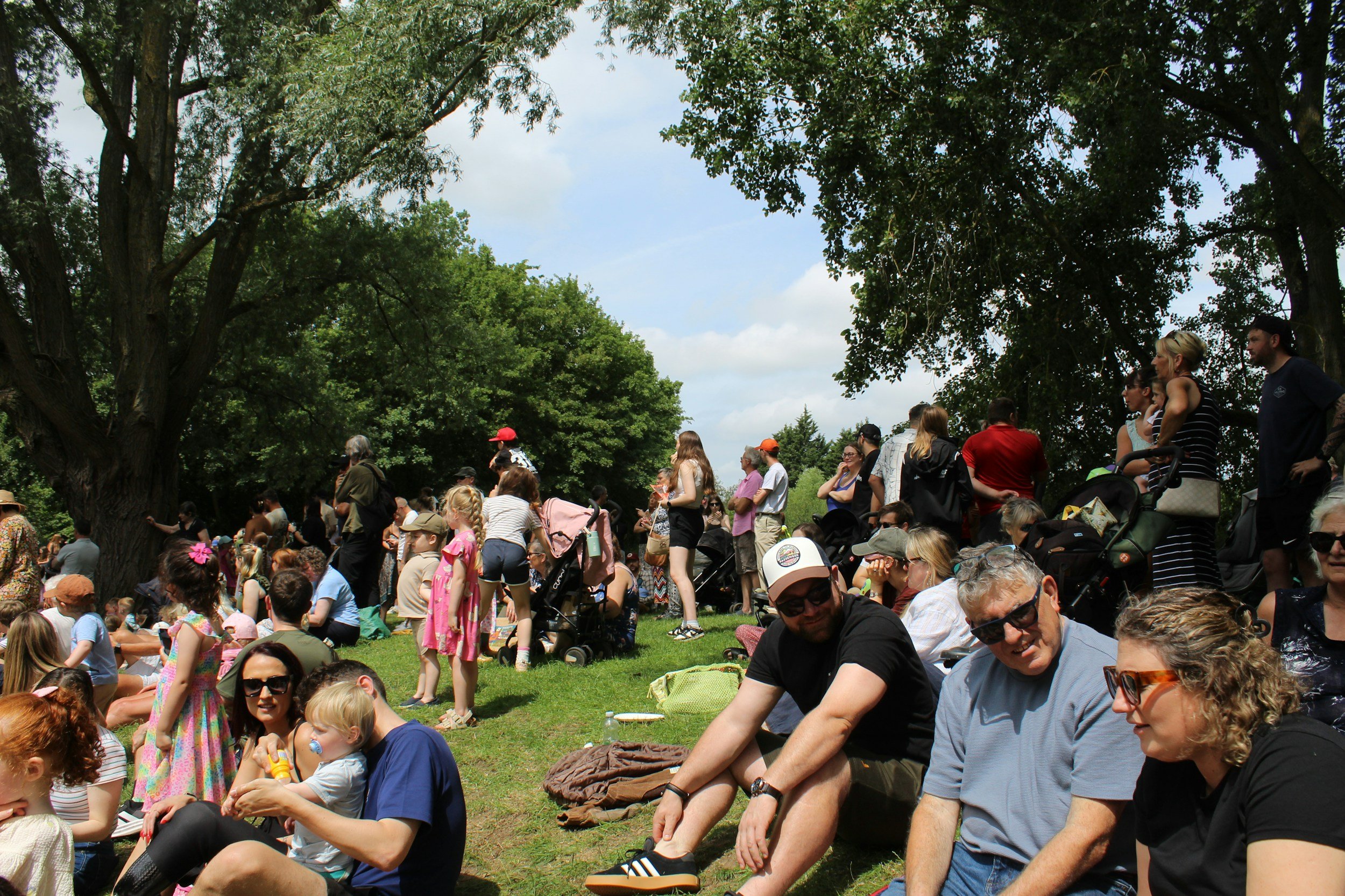 A large crowd of people gathered outdoors under trees, some sitting on the grass and others standing, with a partly cloudy sky in the background.