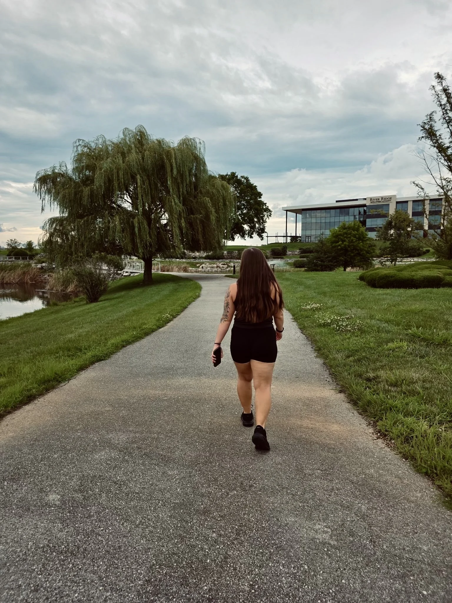 Woman with long hair wearing black shorts and tank top walking on a paved path through a park with green grass, trees, and a modern glass building in the background. Brittany Whitt, NASM Certified Nutrition and Wellness Coach helping women build a he