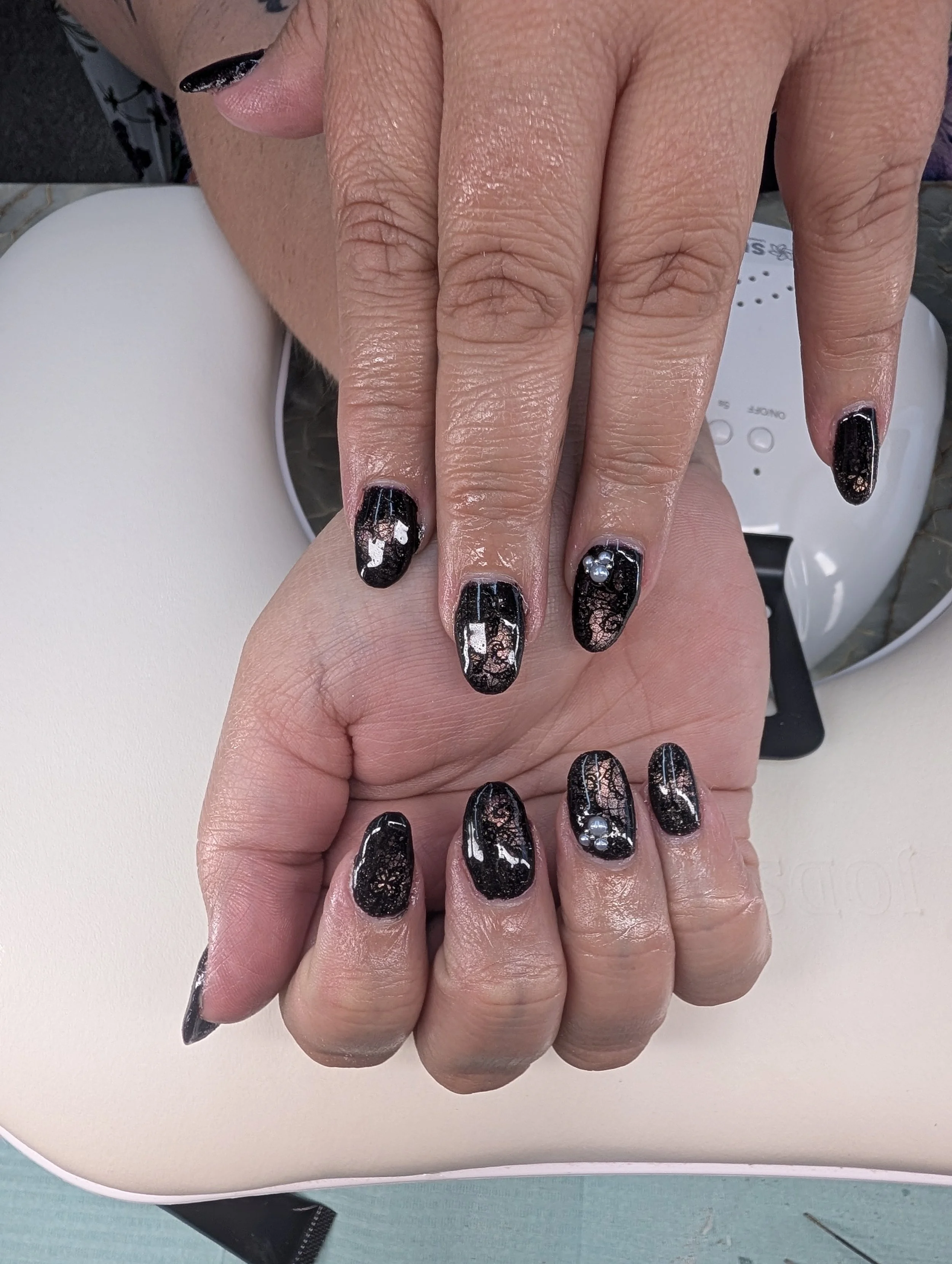 Close-up of hands with freshly painted black nails featuring a lace pattern, rhinestones, and glitter accents, resting on a white surface with a nail dryer in the background.