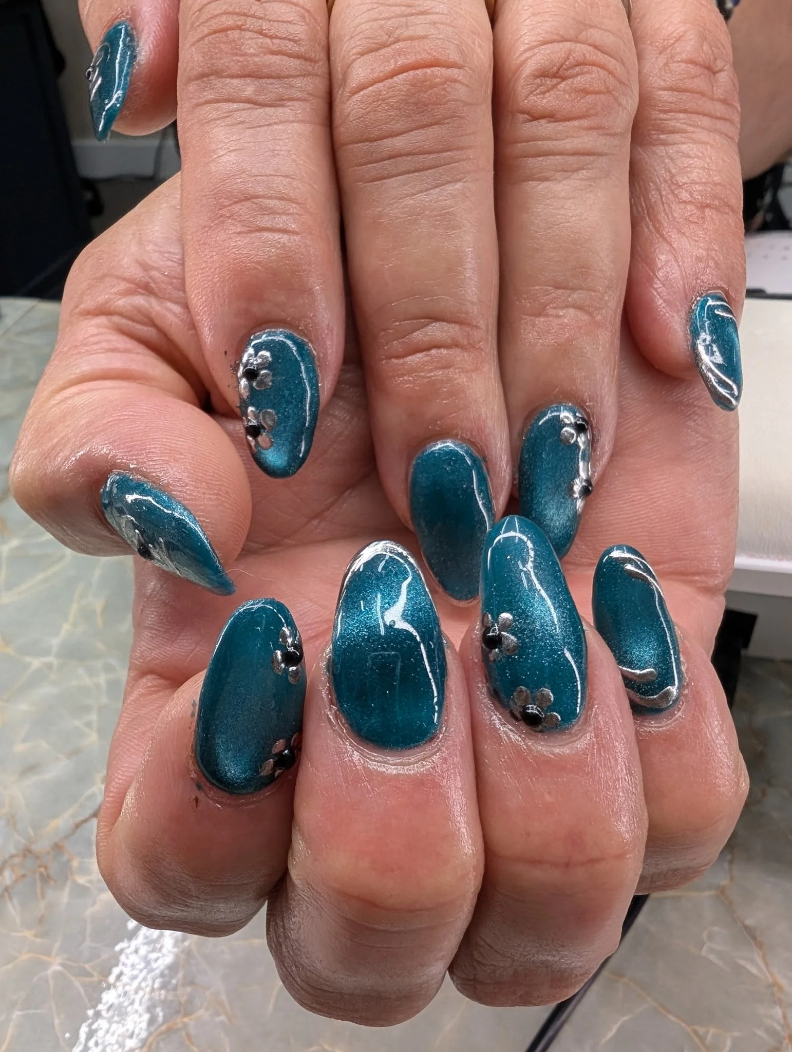 Close-up of a person's hand with long, blue, metallic gel nails, decorated with silver rhinestones and white swirl details.