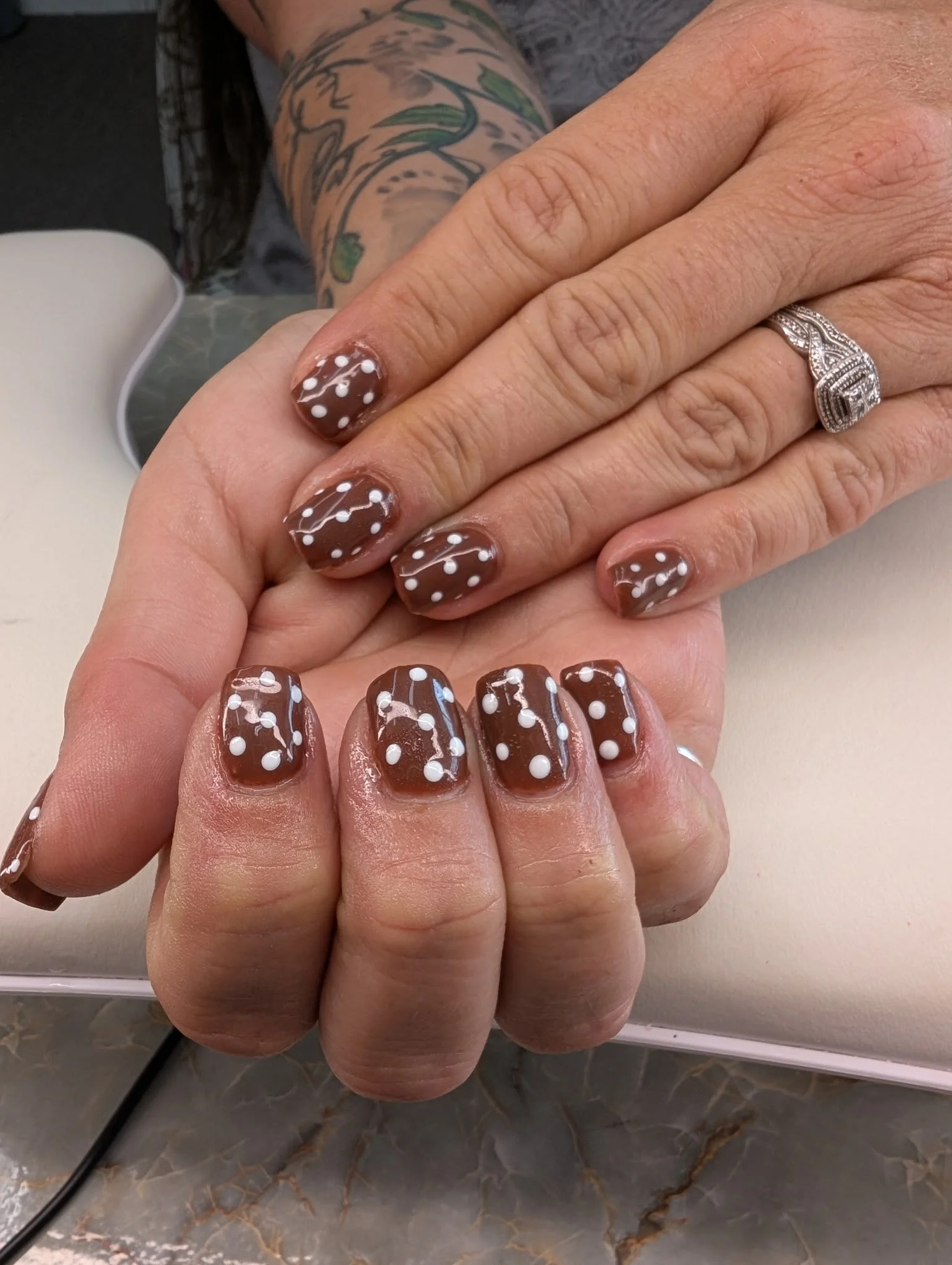 Close-up of a woman's hand with a diamond ring, showing manicured nails painted brown with white polka dots and a white swirl design, resting on another hand with similar nail polish, on a marble surface with part of a tattooed arm visible.