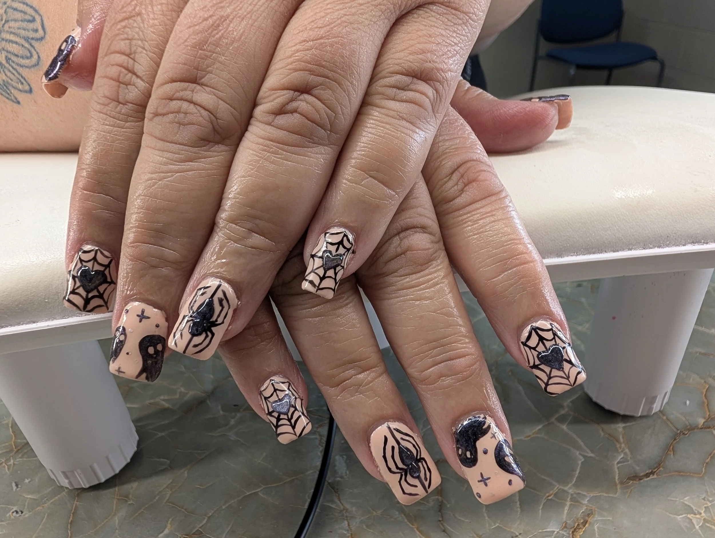 Close-up of a person's hand with Halloween-themed nail art featuring black spiders, cobwebs, bats, and stars on a nude base. The person has a tattoo on their wrist and is resting their hand on a white surface.