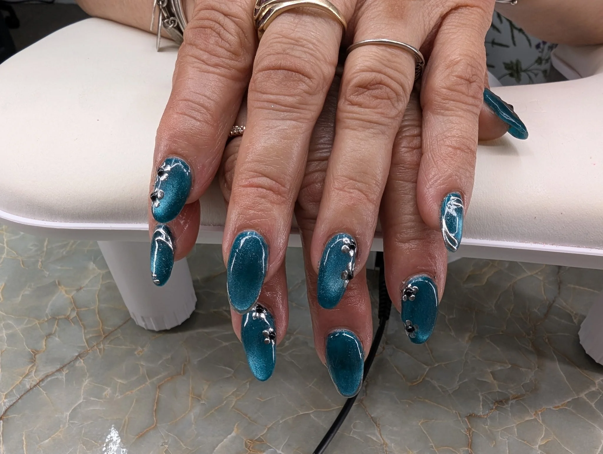 A person with well-manicured nails painted in teal with silver embellishments, wearing multiple rings on their fingers, resting hands on a white cushion in a nail salon setting.