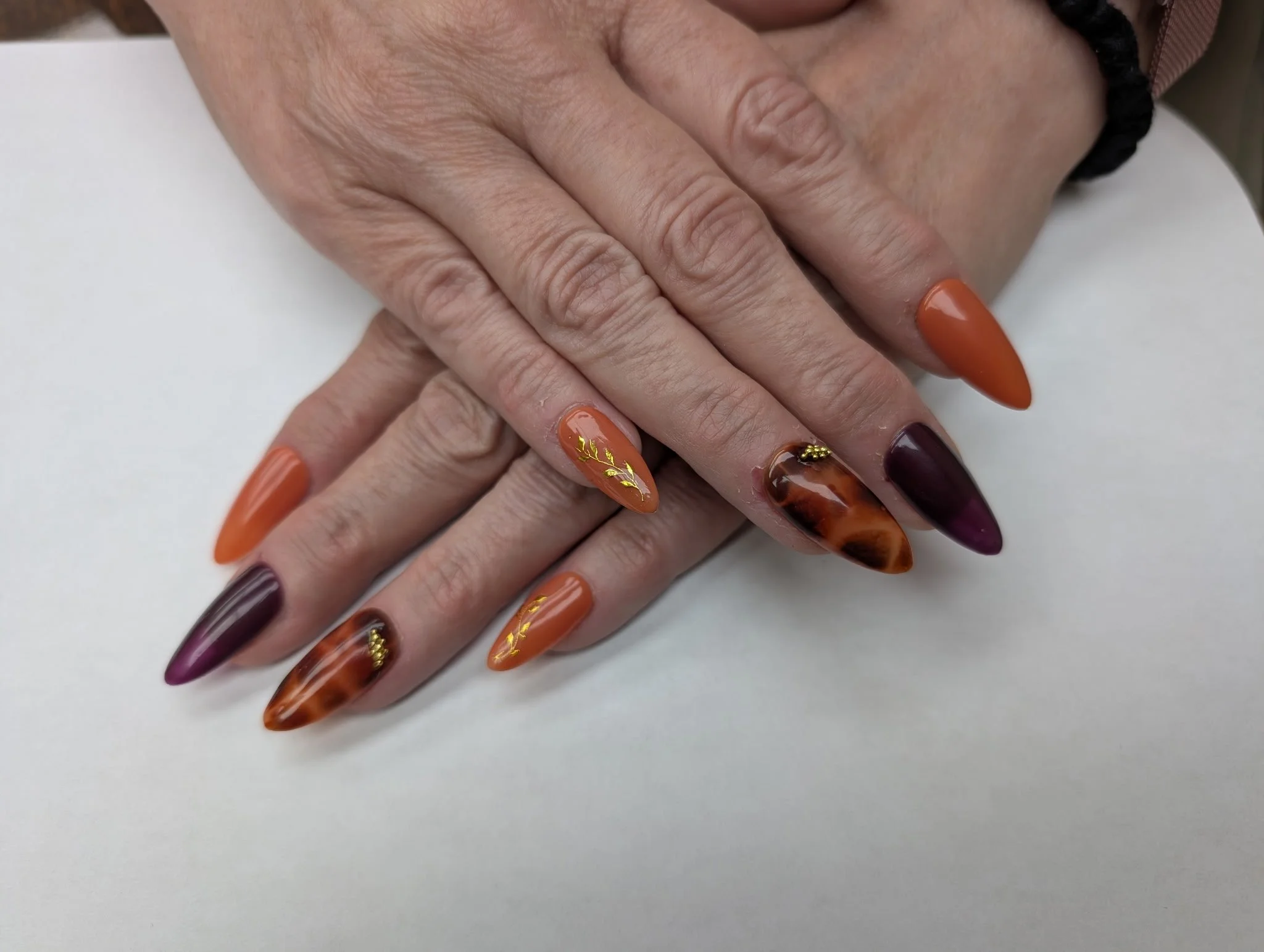 Close-up of a hand with long, decorated nails painted in shades of orange, brown, and maroon, with gold leaf accents.
