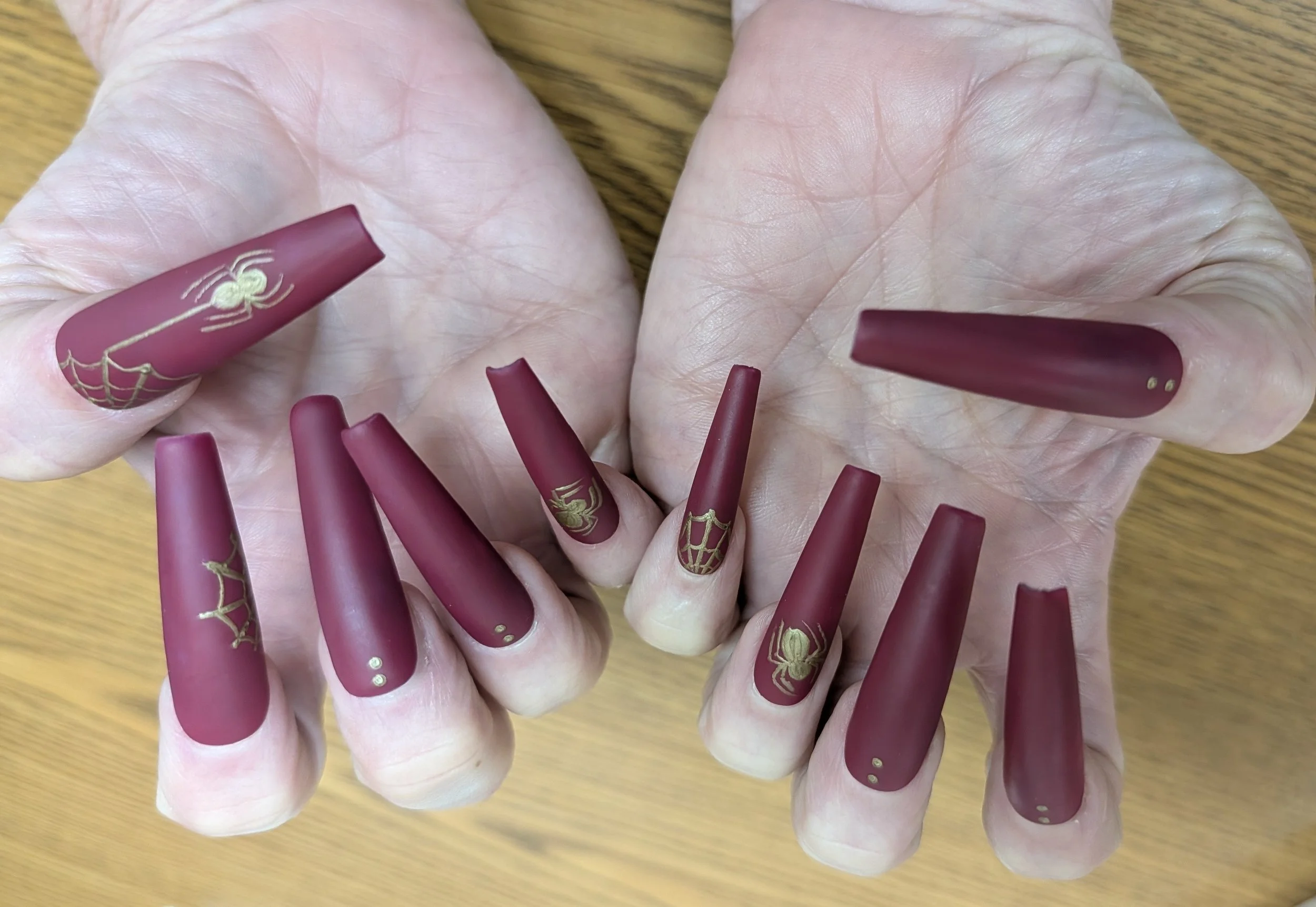 Hand with long matte burgundy nails featuring gold spiderweb design and small gold dots.