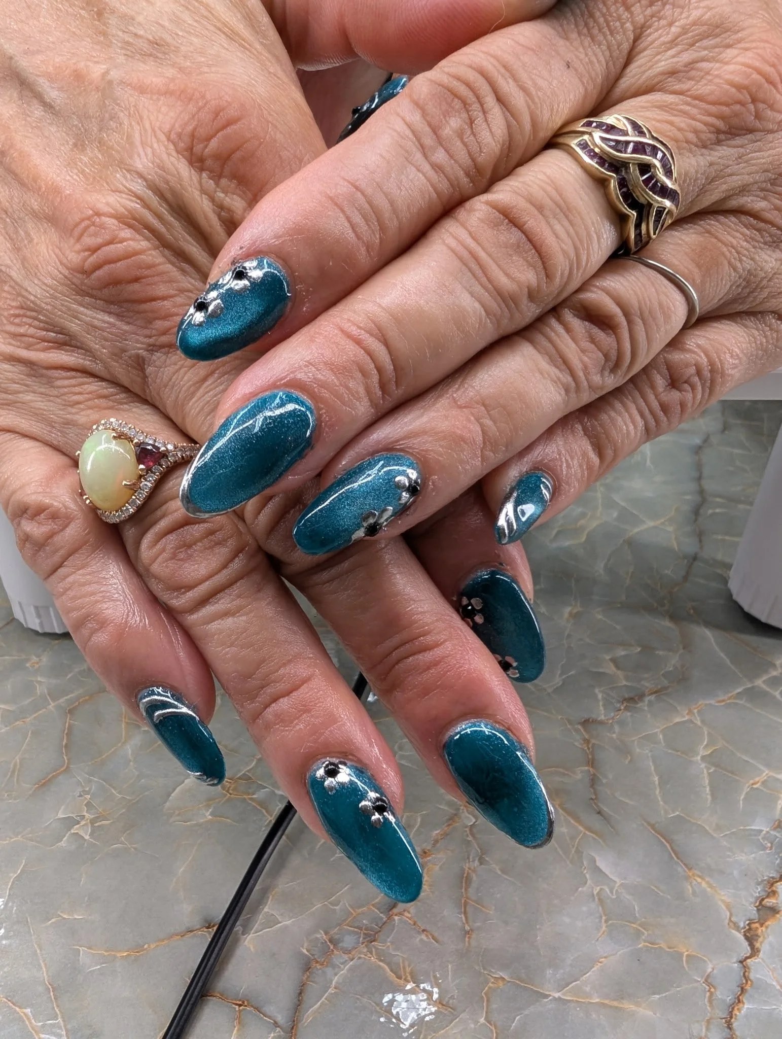 Close-up of hands with manicured nails featuring teal polish, silver accents, and decorative stones. The person is wearing multiple rings, including a gold ring with purple stones and a delicate silver band. The background appears to be a marble surf
