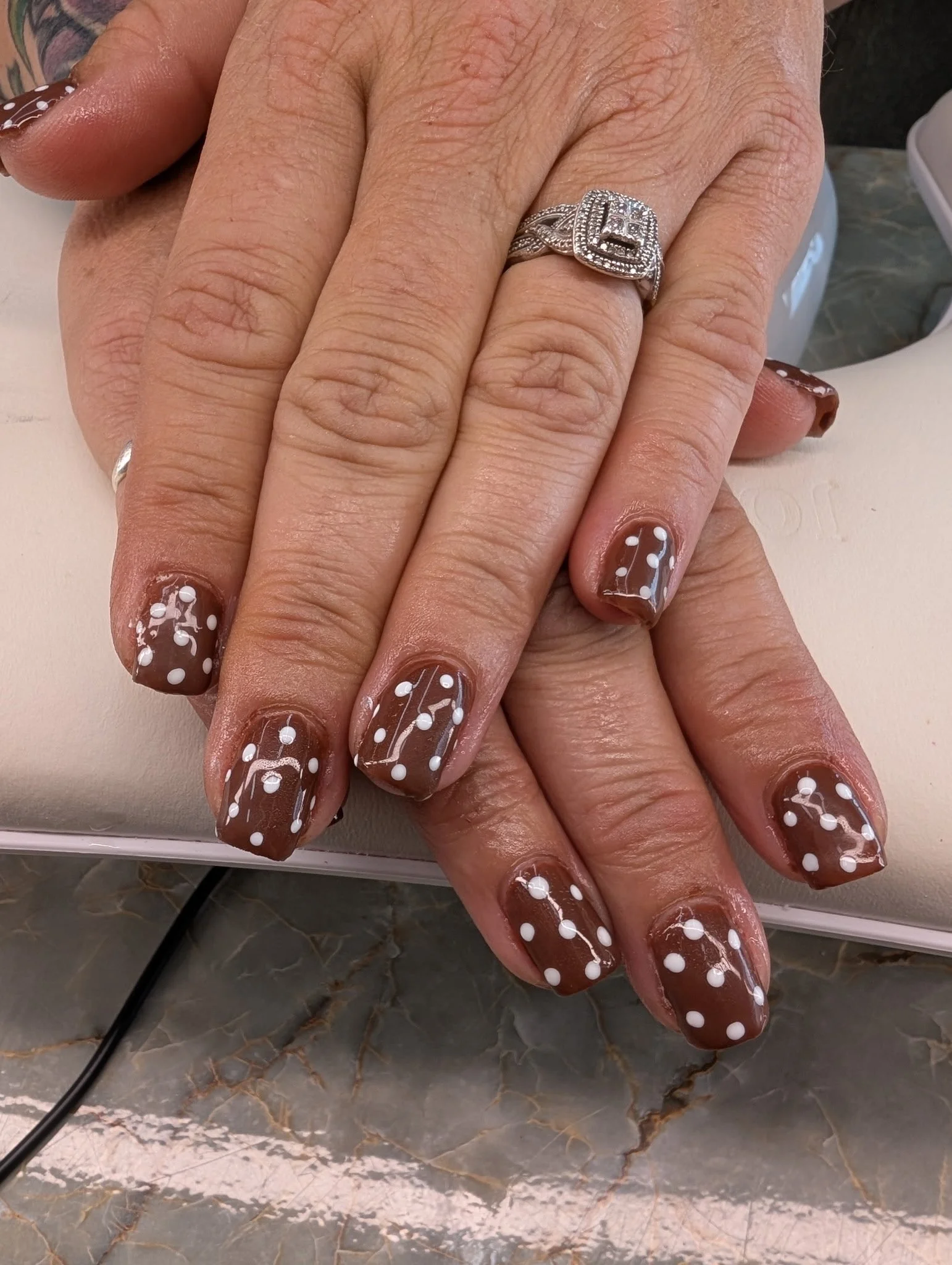 Close-up of a person's hand with manicured nails featuring brown polish and white polka dots, wearing a silver ring with a square design and embedded stones
