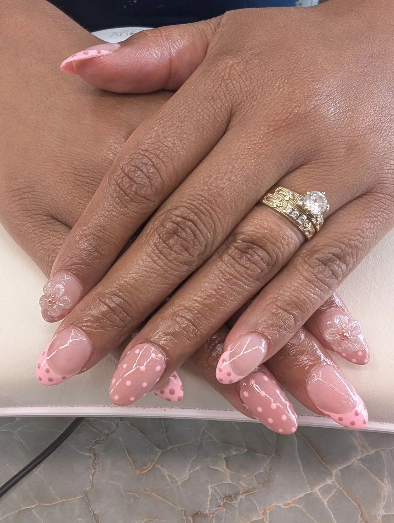 Close-up of a woman's hand with decorated nails, wearing two rings, one with a large center stone. The nails are painted pink with white polka dots and marbled designs.