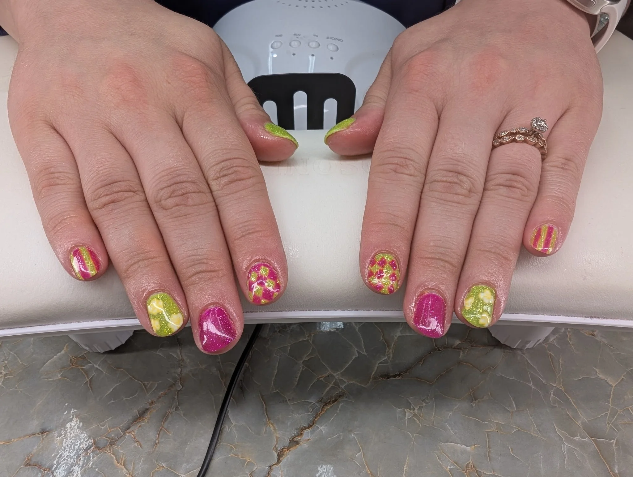 Hands with colorful floral and striped nail art resting on a surface with a nail dryer in the background.