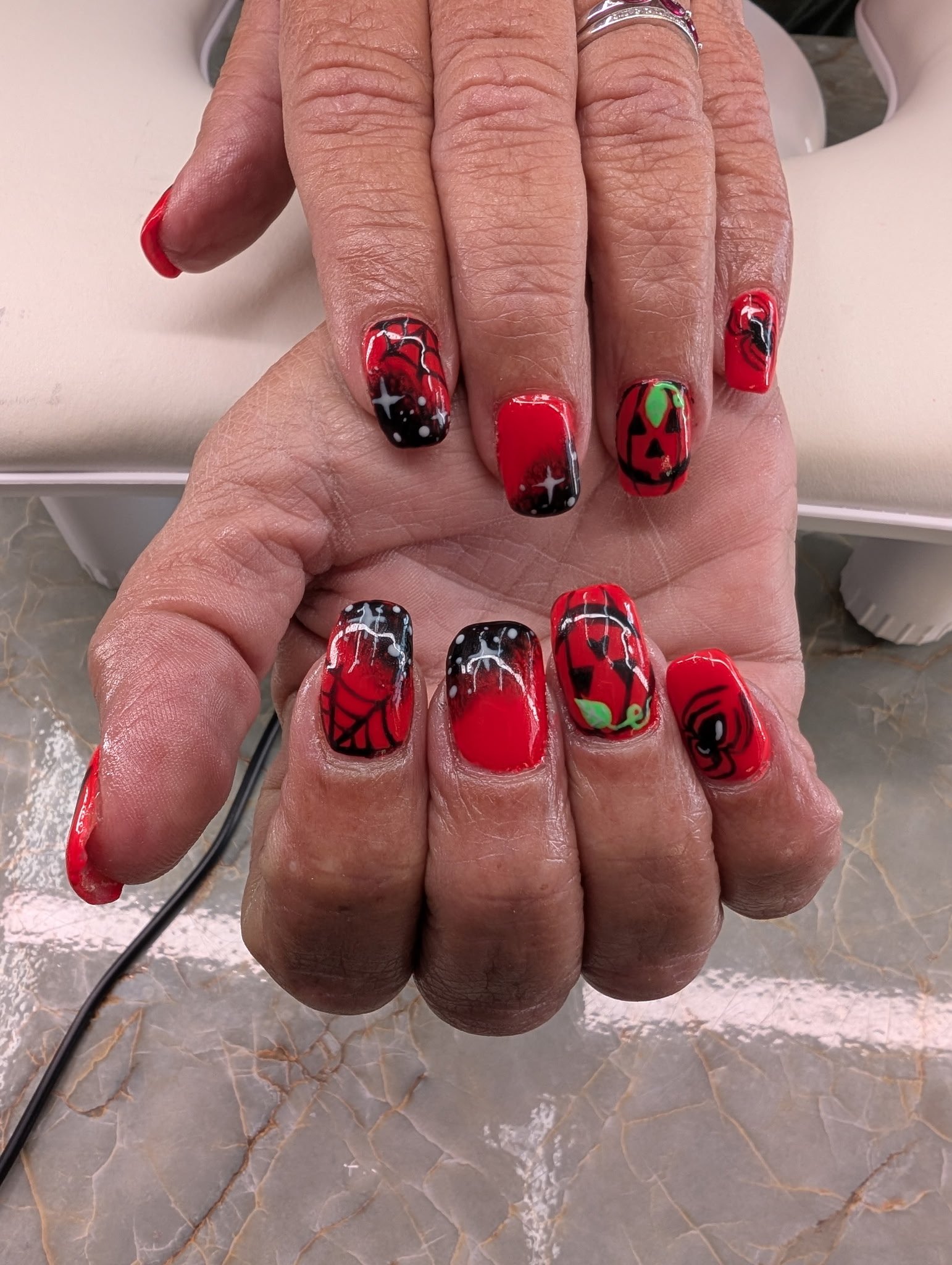 Close-up of a woman's hand with manicured nails featuring Halloween-themed nail art in red, black, white, and green, including a pumpkin, spider web, and ghost designs. The woman is wearing rings on her fingers and is resting her hand on a surface wi