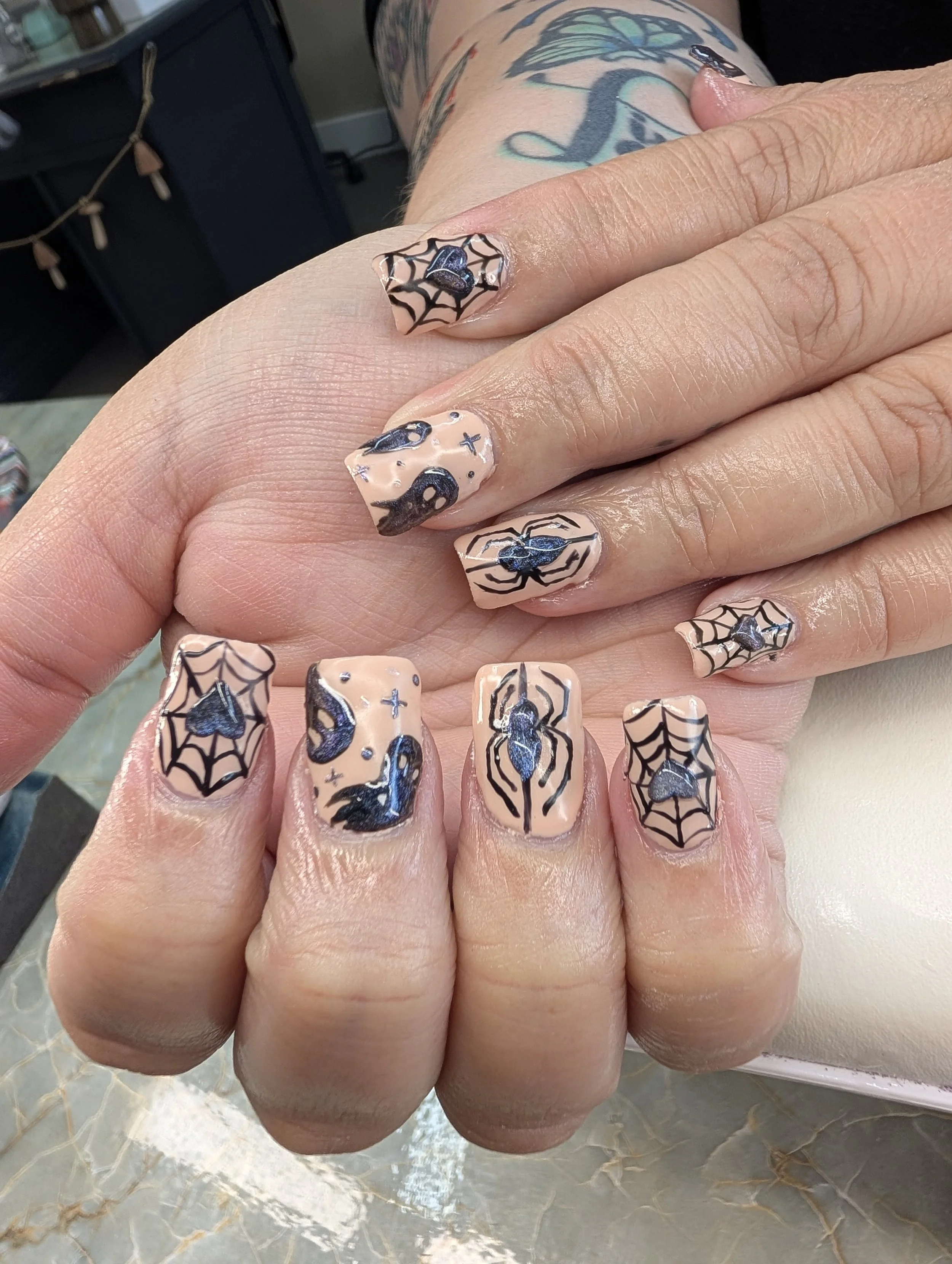 Close-up of a hand with decorated Halloween-themed nails featuring black spiderwebs, spiders, bats, and skulls on a nude base color.