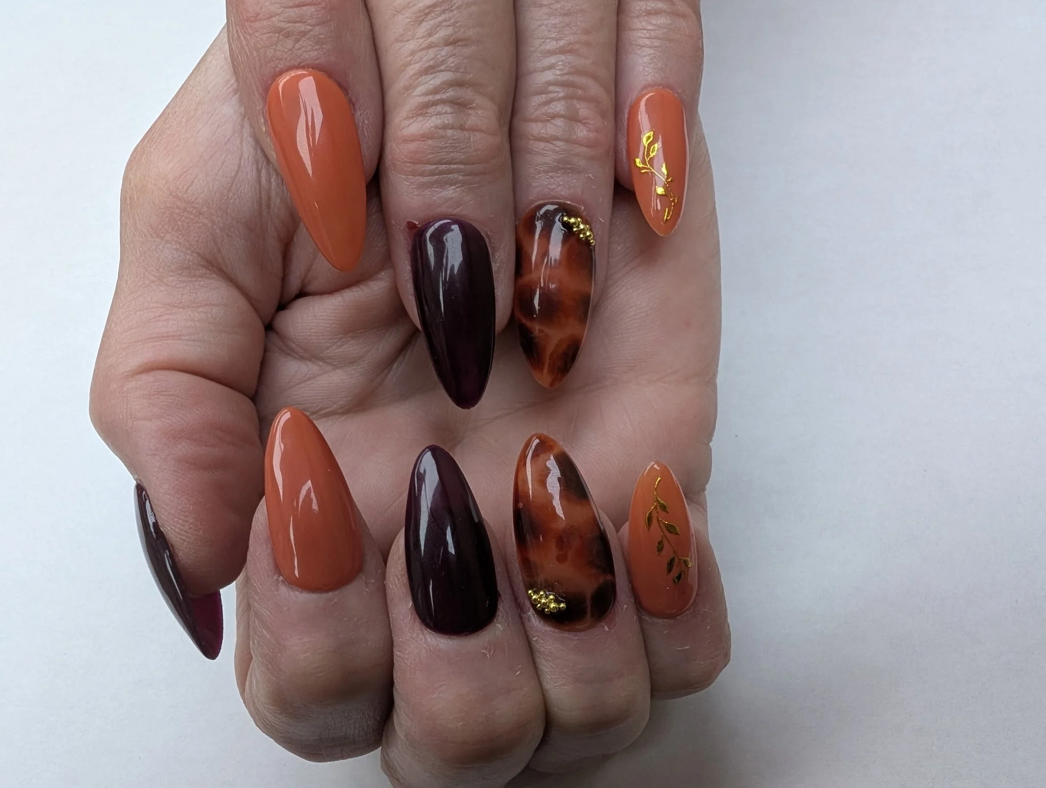Hand with long, almond-shaped nails painted in various shades of orange, brown, and tortoiseshell, some with gold leaf accents and small gold beads