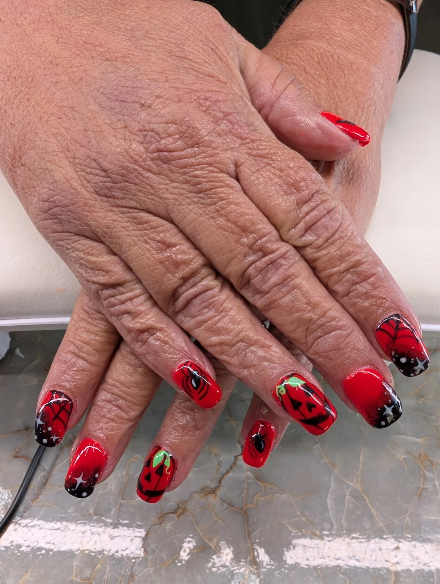 Close-up view of two hands with Halloween-themed nail art, featuring red, black, green, and white colors, with designs like spiders, pumpkins, and stars, resting on a marble surface.