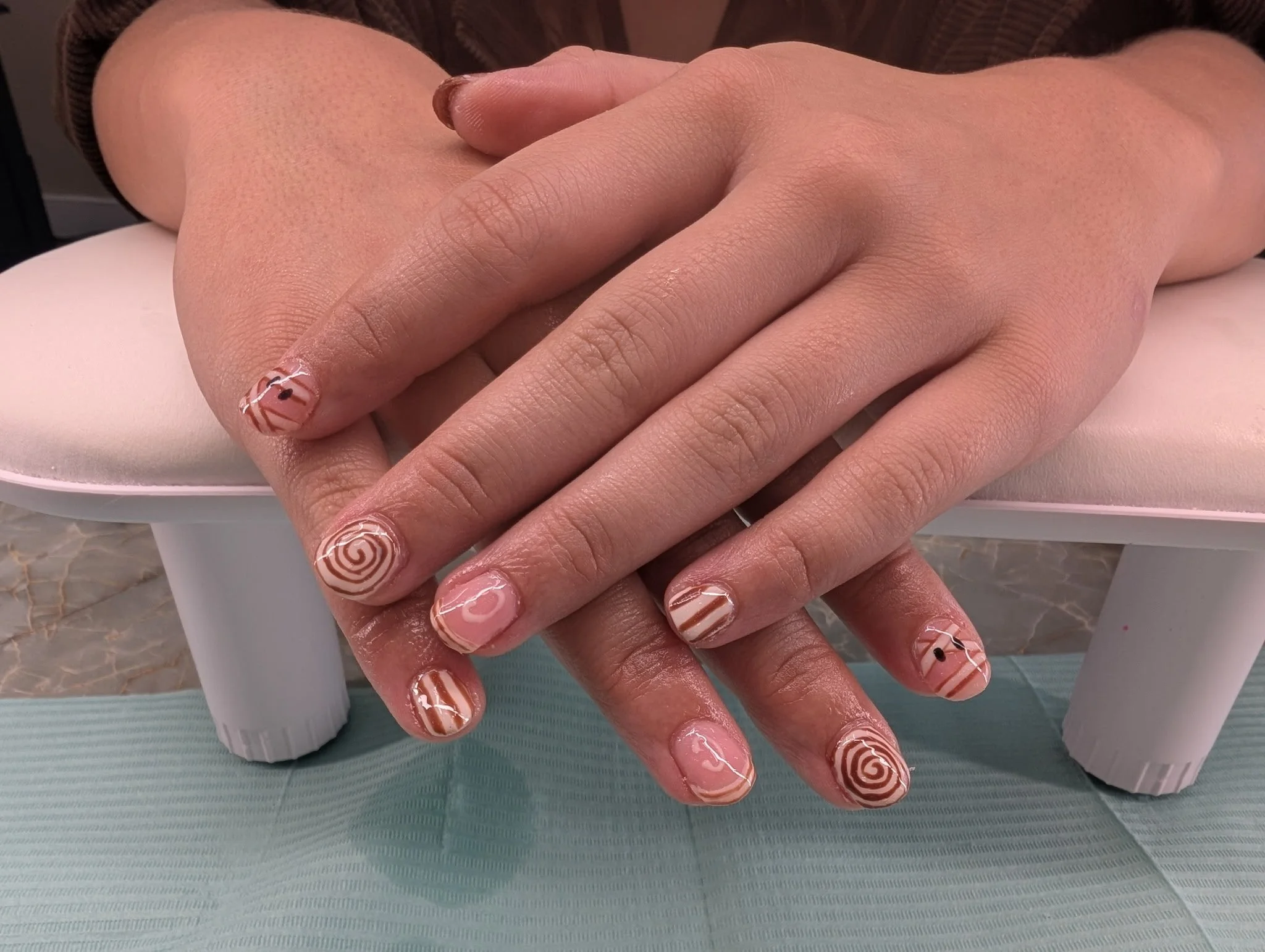 Close-up of two hands with decorated nails resting on a surface. The nails have tan base color with white swirl and stripe designs.