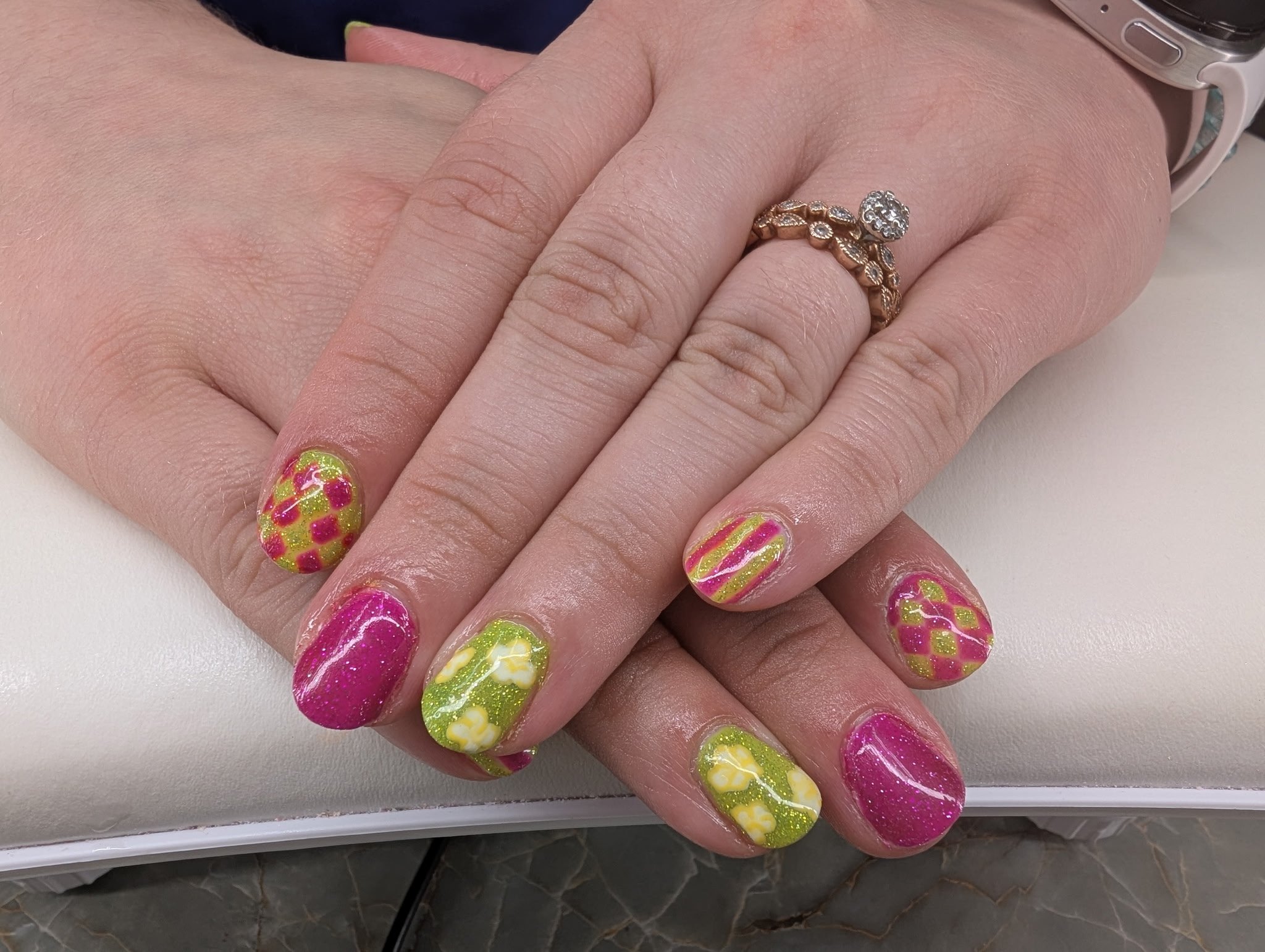 Close-up of a person's hand showing colorful nail art with a pink, yellow, and green pattern, and a gold ring on the ring finger. A silver watch is on the wrist in the background.