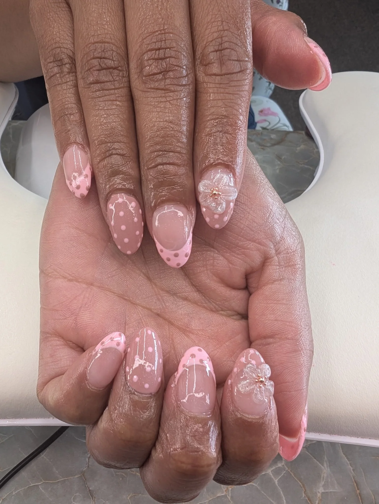 Close-up of a person's hand with manicured nails, featuring a pink base and white polka dot design on some nails, with a jewelry ring on the ring finger. The hand is resting on a white table with a decorative mirror or reflective surface in the backg