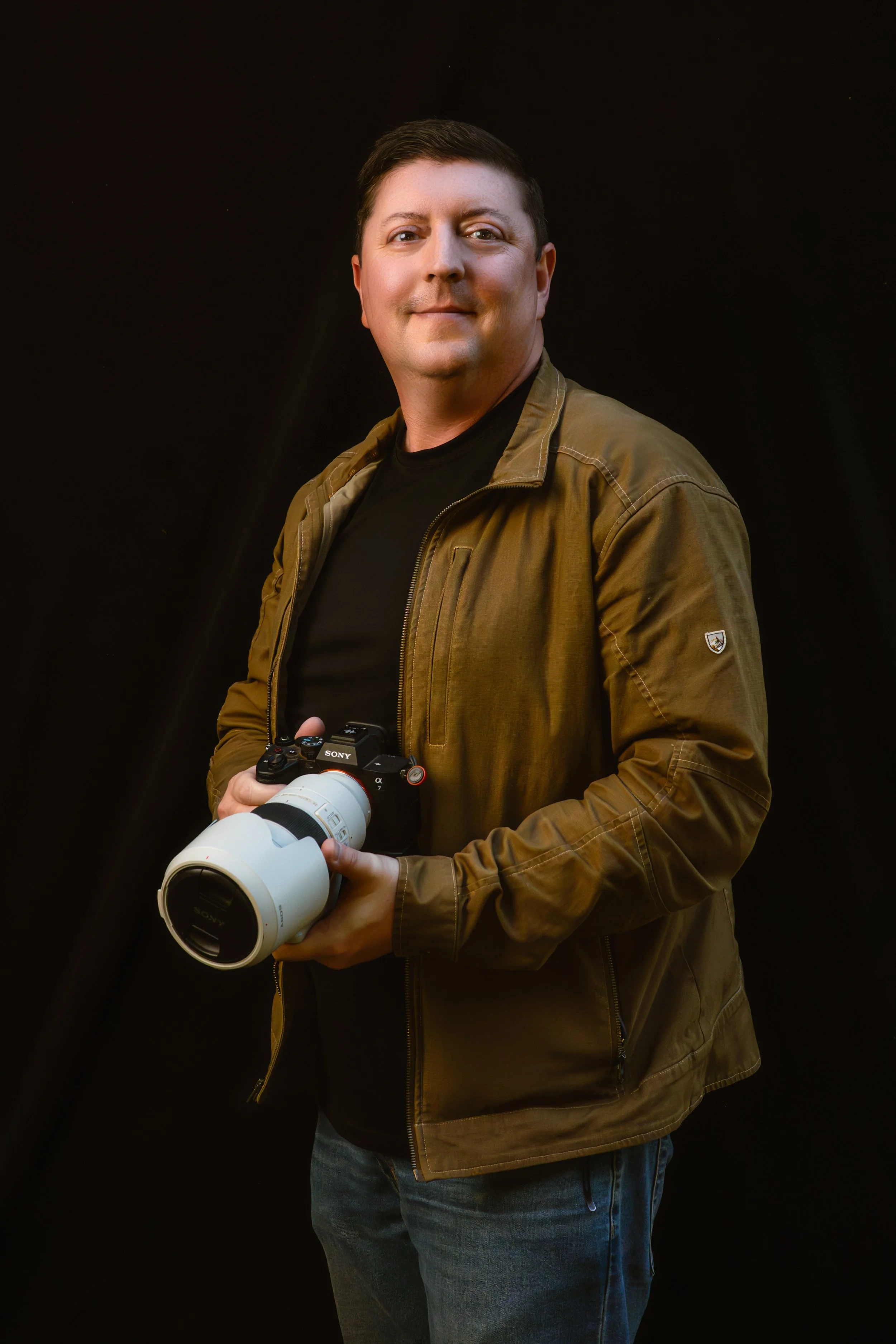 A man holding a Sony camera with a large white telephoto lens, wearing a brown jacket and black shirt, standing against a black background.