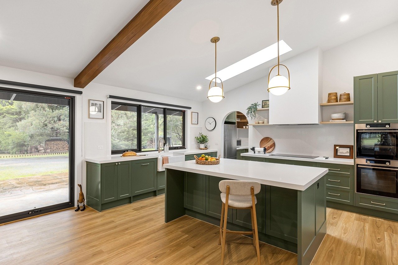 Bright modern kitchen with green cabinetry, white countertops, and large windows, featuring a kitchen island with a fruit bowl, wood flooring, pendant lighting, and a view of trees outside.