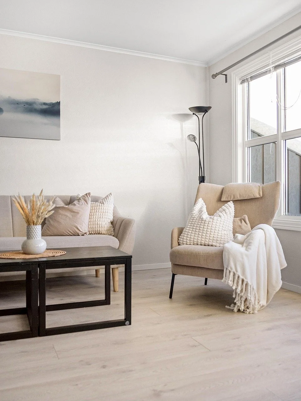 A cozy living room with beige armchair, beige sofa, black coffee table, white vase with dried plants, and a floor lamp near a window with blinds.