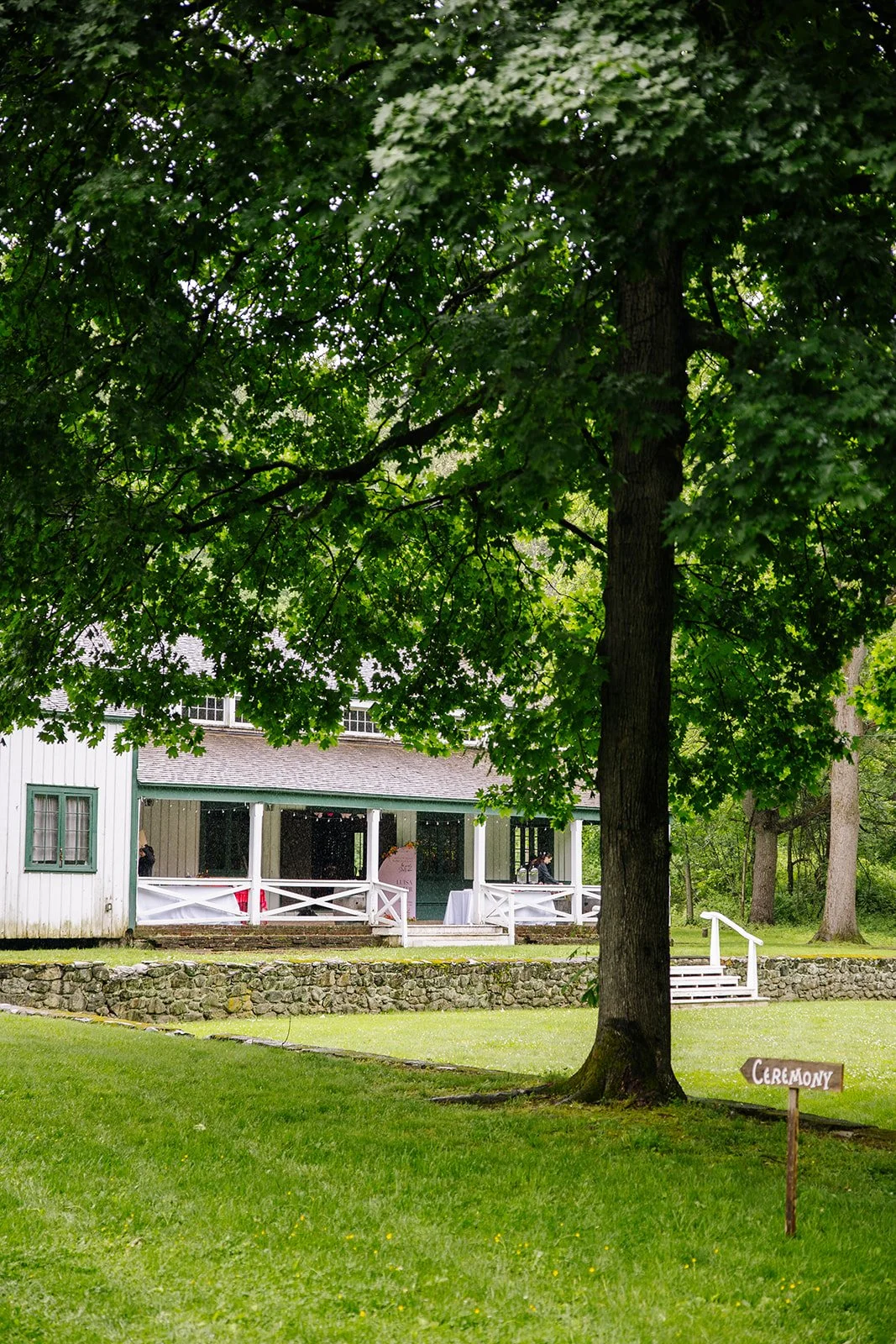 A large tree with green leaves in front of a white building with a porch. A small wooden sign with the word "CEREMONY" points to the right, indicating an outdoor wedding setup.