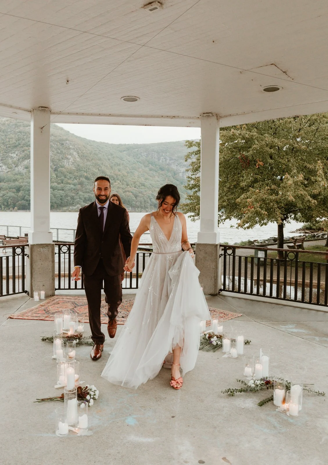 A bride and groom walking hand in hand outdoors on a gazebo with candles and flowers, with a lake and mountain scenery in the background.