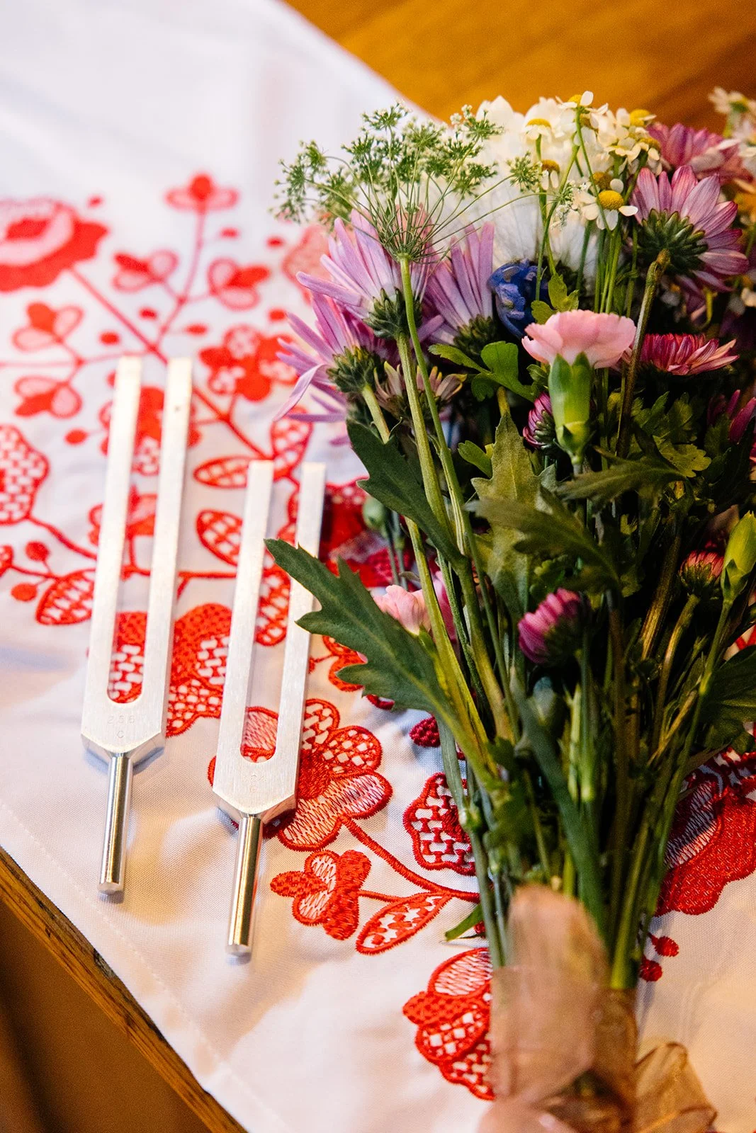 A bouquet of mixed flowers, including pink, purple, white, and yellow blossoms, beside a white embroidered tablecloth with red floral patterns. There are two white candles on the table.