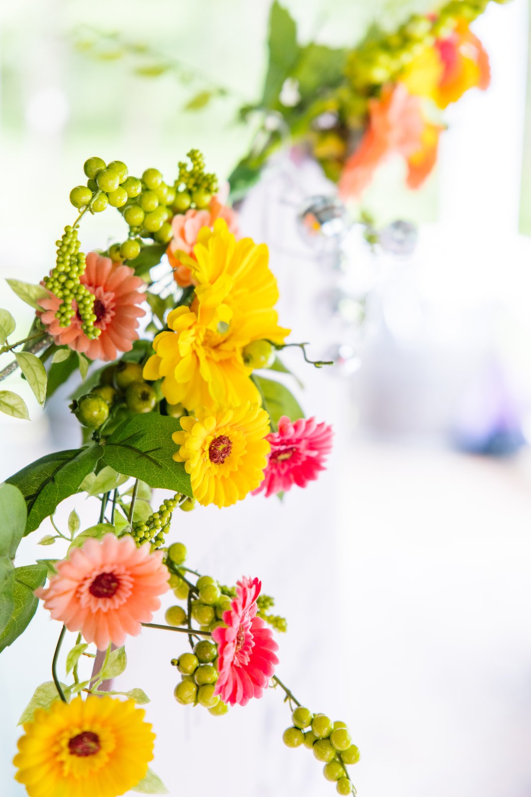 A colorful flower arrangement with yellow, pink, and orange flowers, accompanied by green leaves and small green berries, set against a blurred bright background.