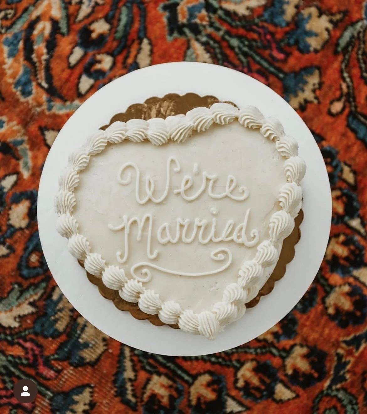 Heart-shaped wedding cake with white frosting and piped border, featuring a message that says "We're Married", placed on a patterned carpet.