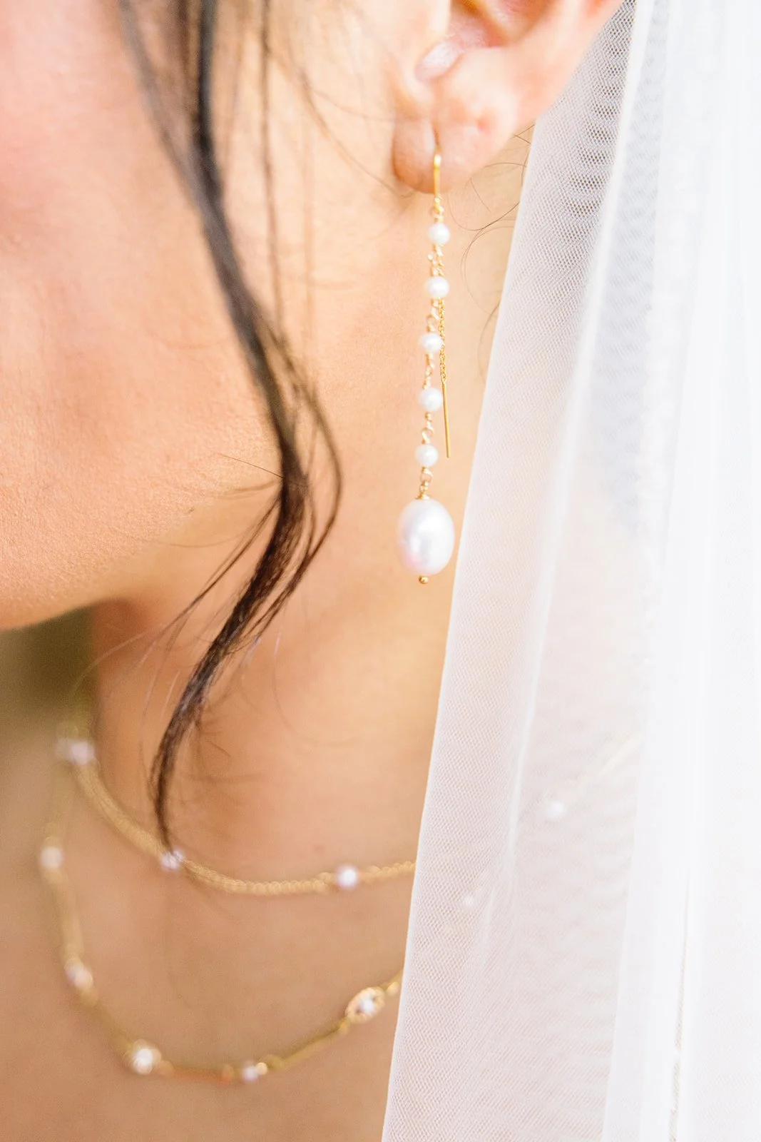 Close-up of a woman wearing pearl jewelry, including a long pearl earring and a matching pearl necklace, with a white veil partially covering her face.