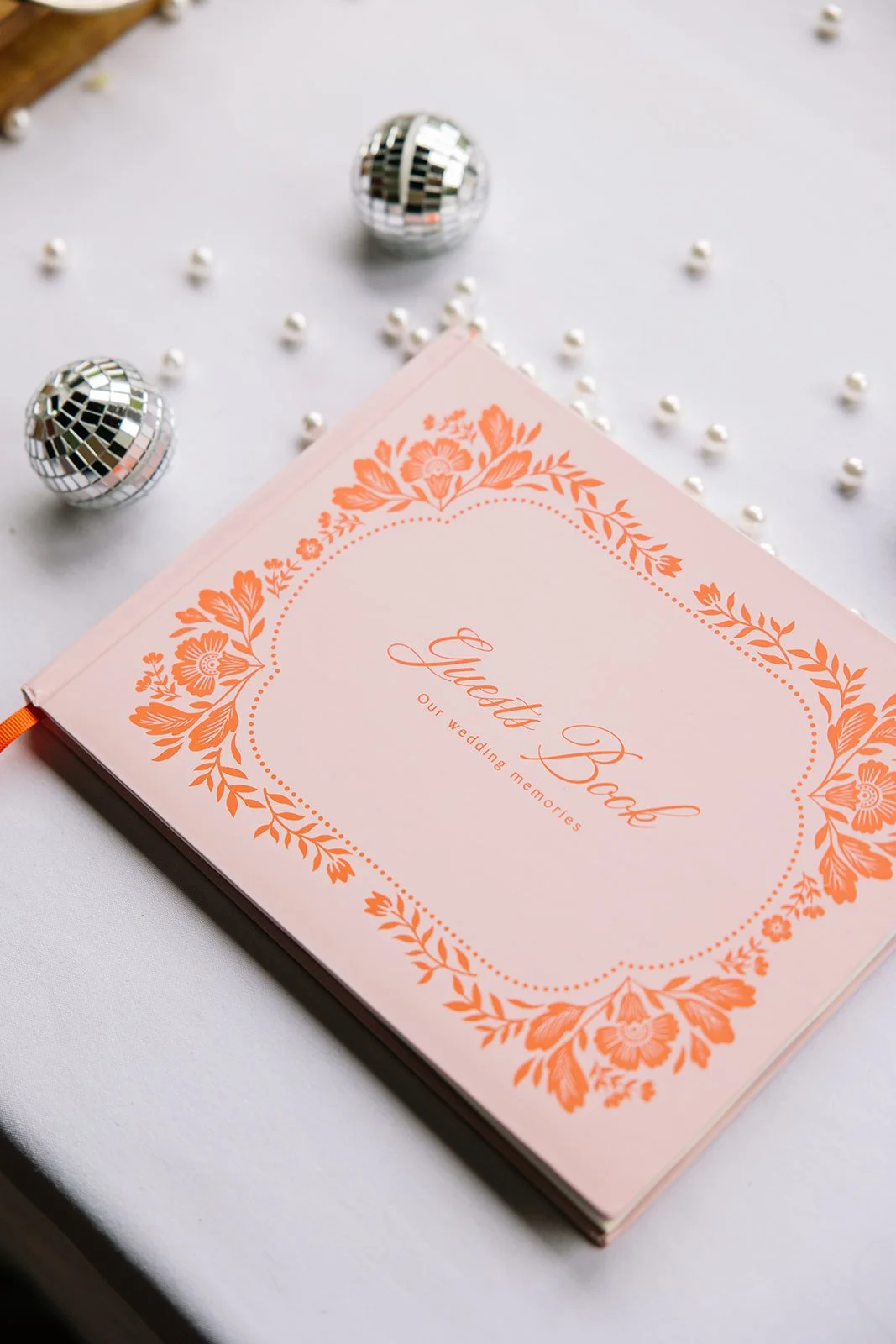 A pink wedding album with orange floral border and script text that reads 'Guest Book Our wedding memories', surrounded by small pearls and disco balls.