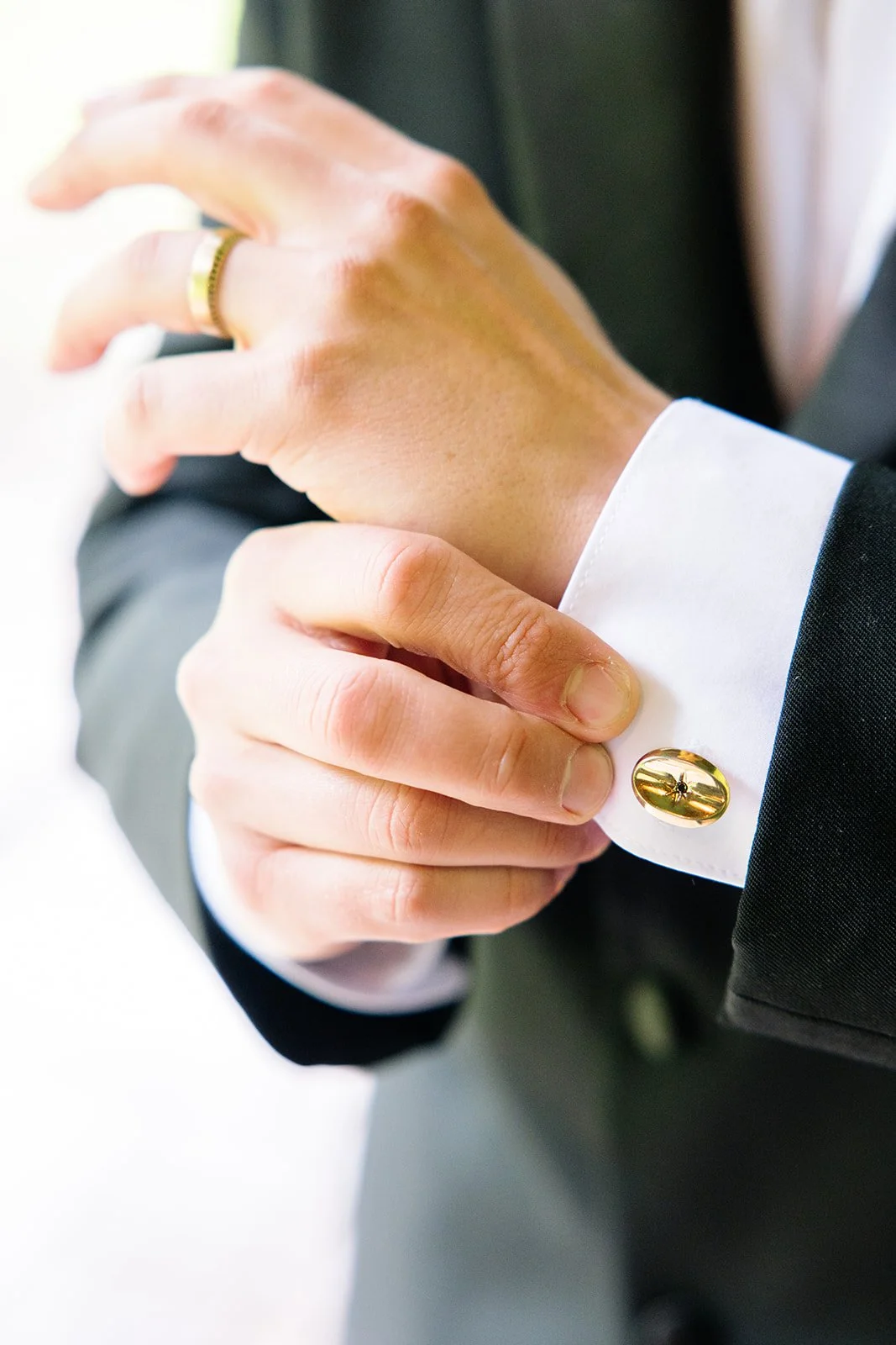 Close-up of a man adjusting his cufflinks on a white shirt cuff, wearing a black suit jacket, with a gold ring on his finger.