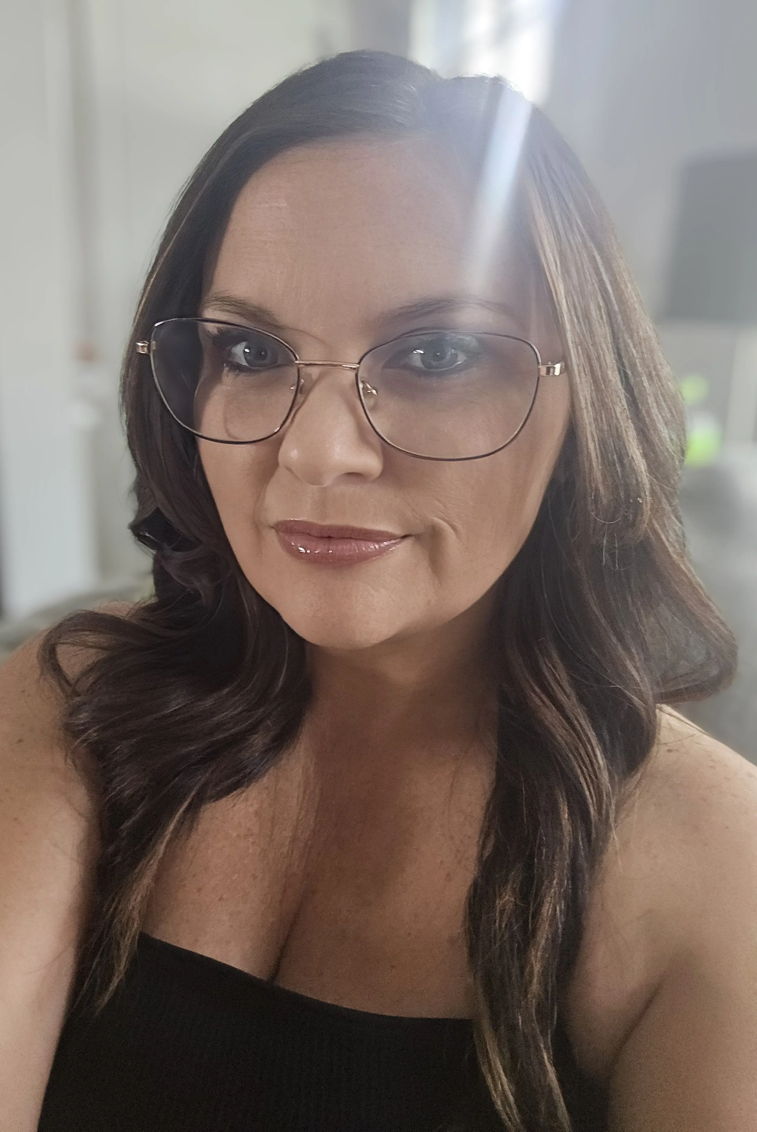 A woman with long dark wavy hair wearing glasses and a black tank top, smiling in a close-up selfie indoors with natural light.