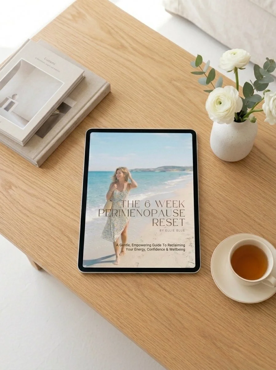 An e-book titled 'The 6 Week Perimenopause Reset' by Ellie Blue resting on a wooden table next to a cup of tea, a vase with white flowers, a stack of books or magazines, and a tablet with a beach scene on the cover.