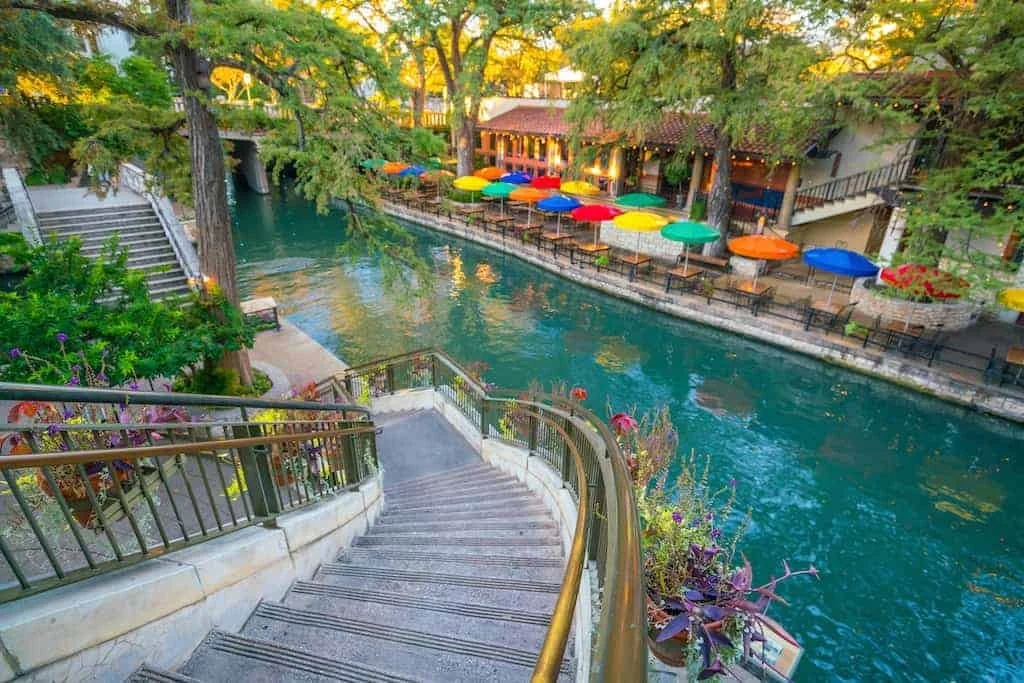 A scenic riverside area with a staircase leading down to the water, surrounded by colorful umbrellas and trees.