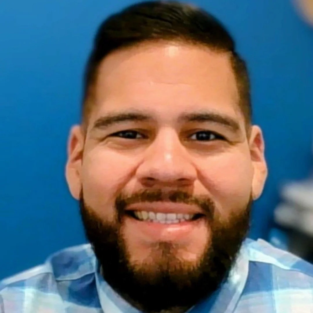 Close-up of a smiling man with short dark hair, a beard, and wearing a light blue plaid shirt against a blue background.