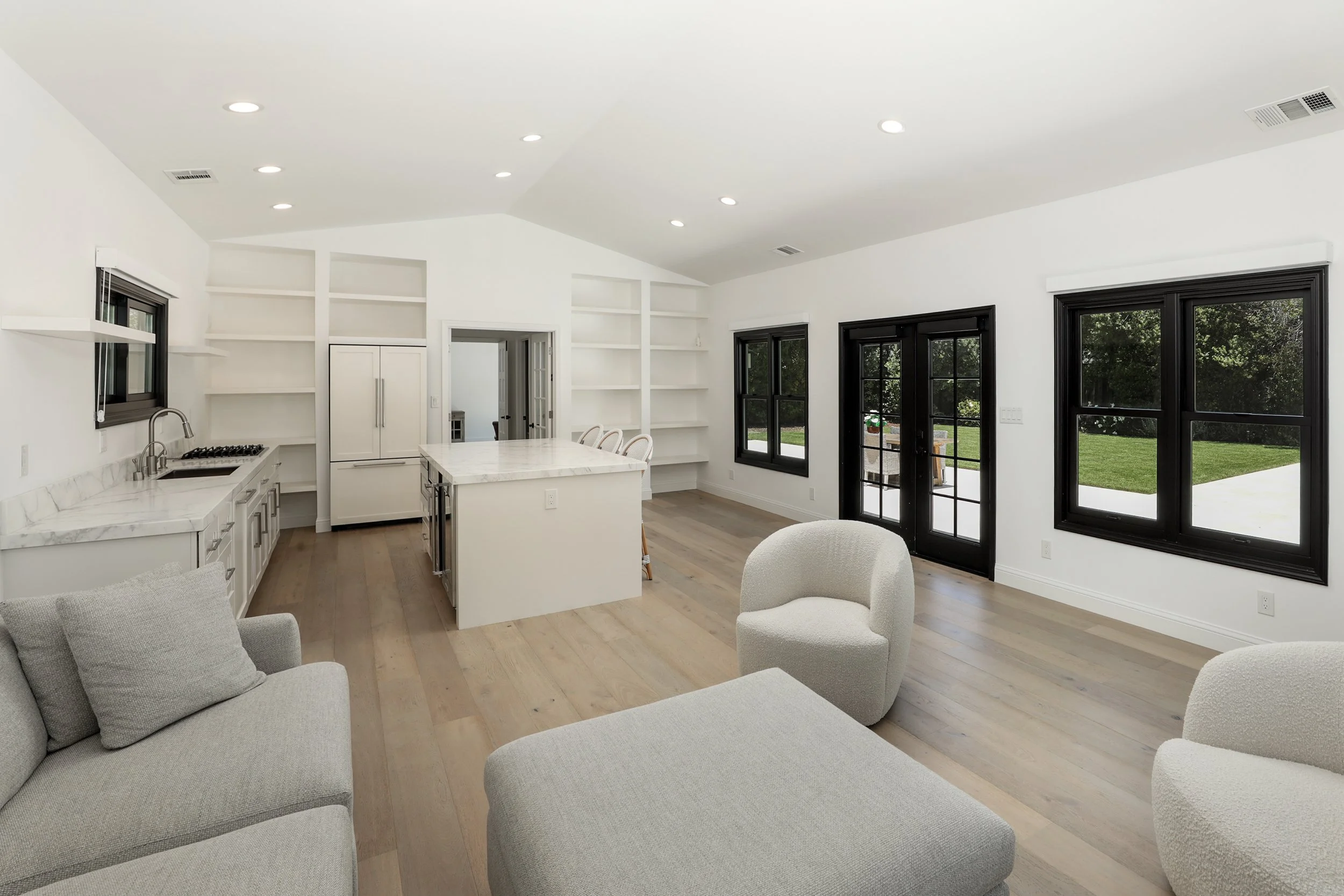 Bright living room with white walls, large windows with black frames, a kitchen with a white marble island, open shelves, and a door leading outside to a lawn.