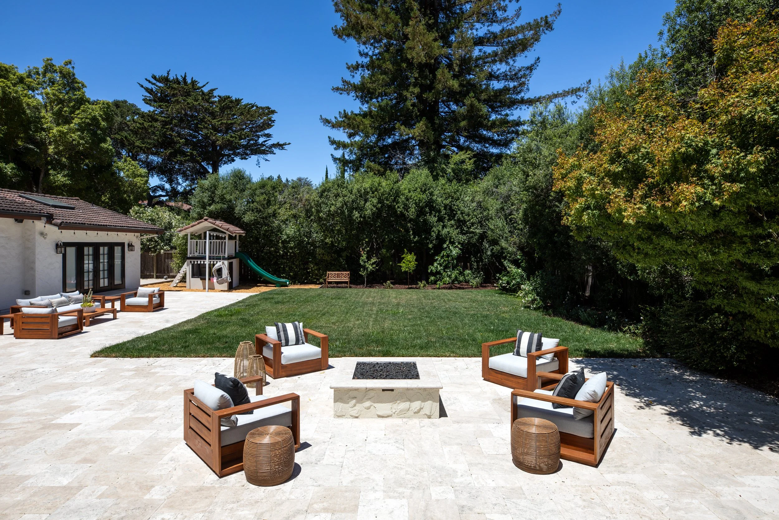 A backyard patio with outdoor seating including white cushioned chairs and small tables. A lawn with a grassy border and a children's playhouse with a slide are visible, surrounded by various trees under a clear blue sky.