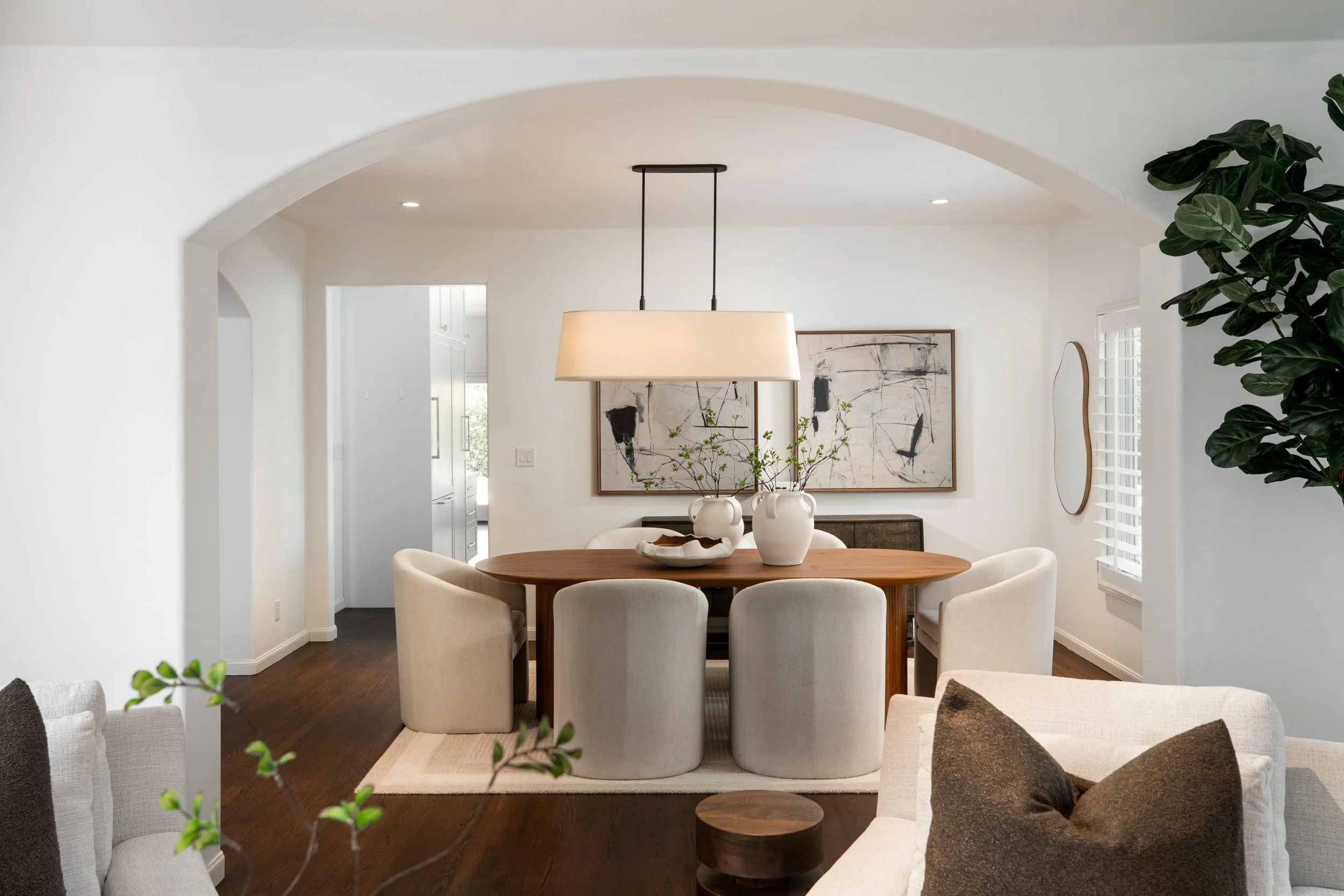 A modern dining room with a round wooden table surrounded by cream upholstered chairs. Above the table hangs a large beige pendant light. The wall features abstract artwork, and there are decorative vases with greenery on the table. A large potted pl