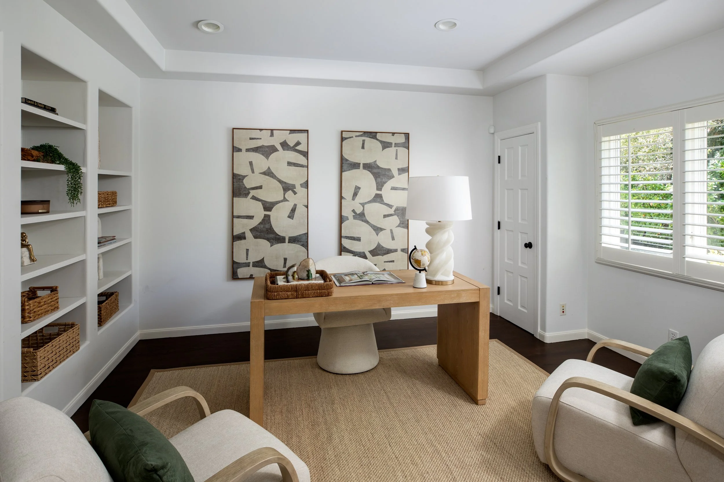 A cozy home office with a wooden desk, beige chair, white walls, framed abstract art, a standing lamp, a bookshelf with baskets, and a window with white blinds.