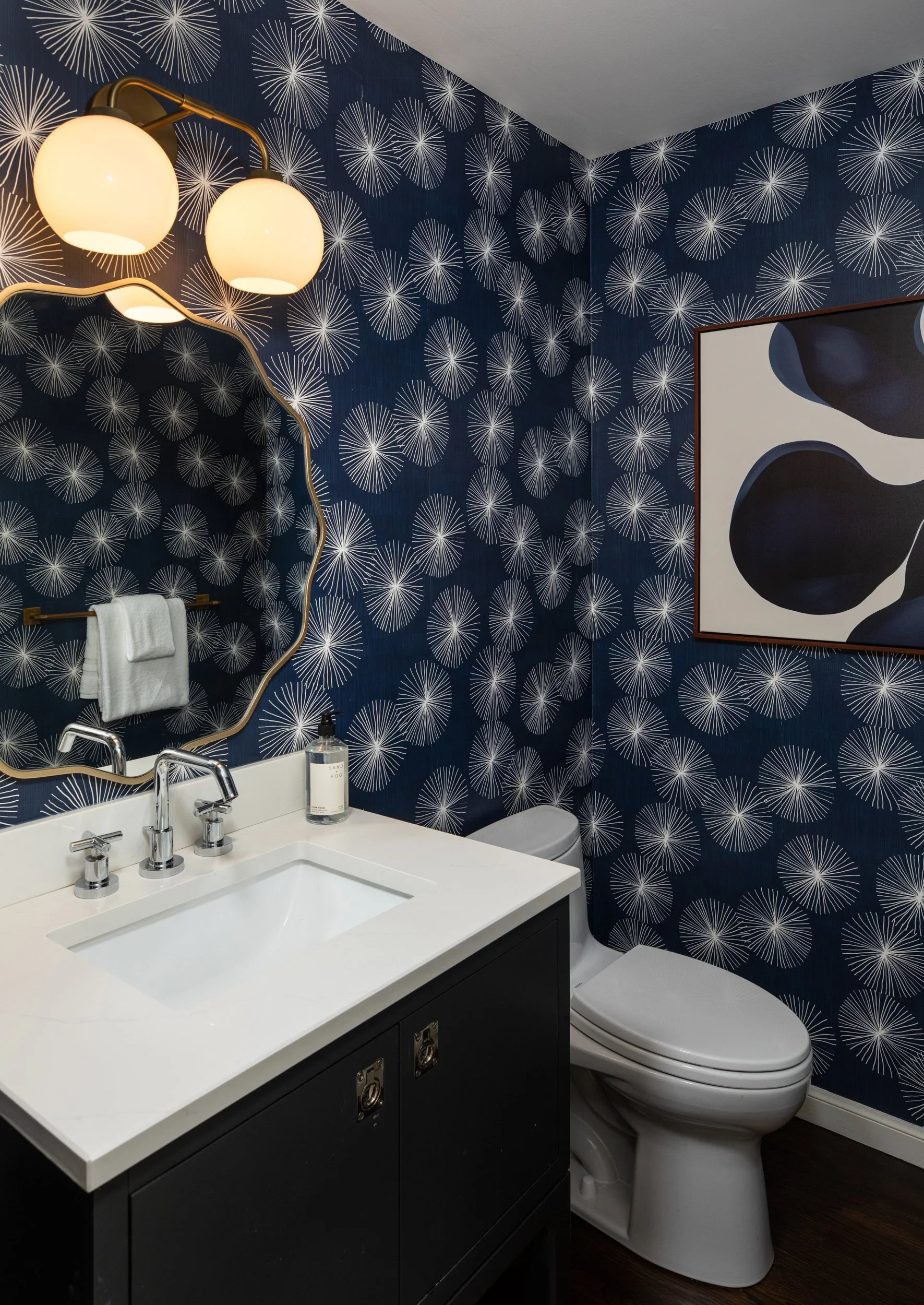 A bathroom with dark blue wallpaper featuring white starburst patterns, a white sink with modern chrome fixtures, a black cabinet underneath, a white toilet, a large mirror with a gold wavy frame, a clear soap dispenser, a towel hanging on a golden b