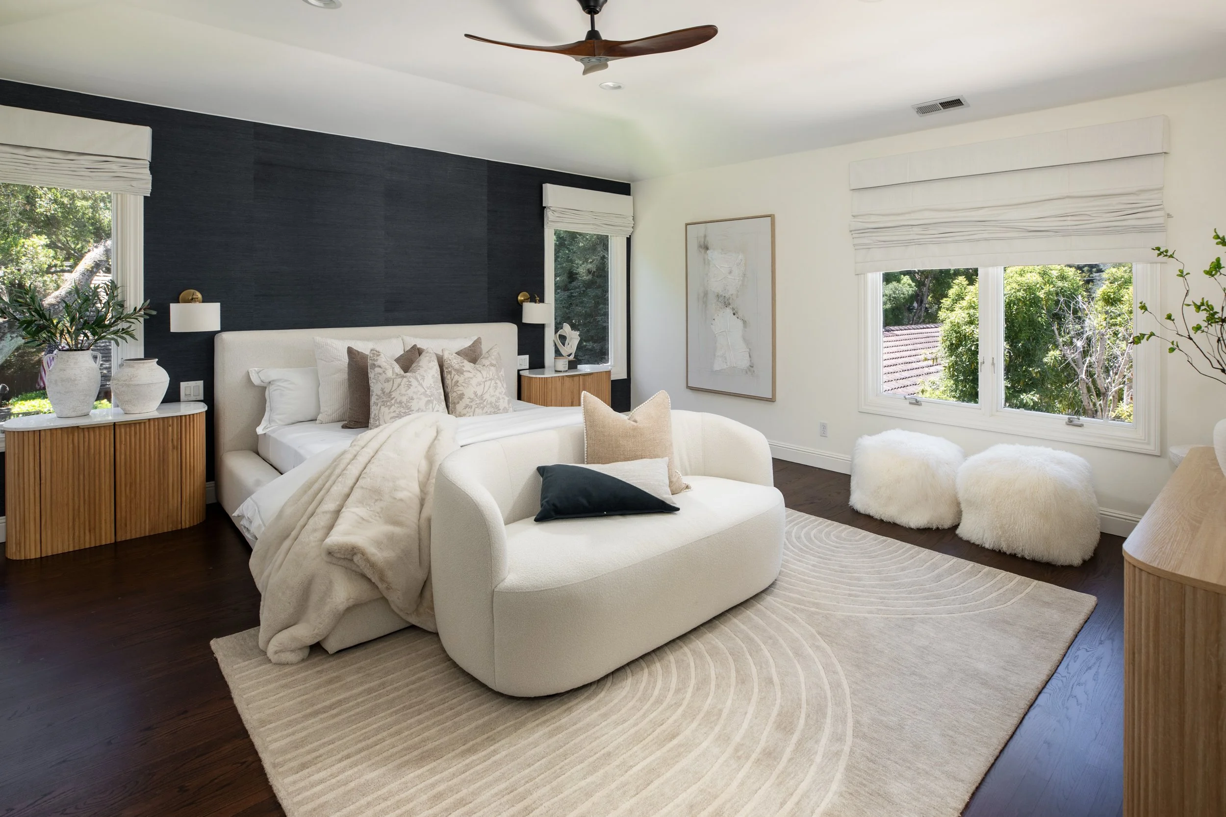 A modern bedroom with a white upholstered bed, beige and patterned pillows, dark wood floor, cream and dark accent wall, large windows with white roman shades, abstract artwork, and plush white and beige furniture.