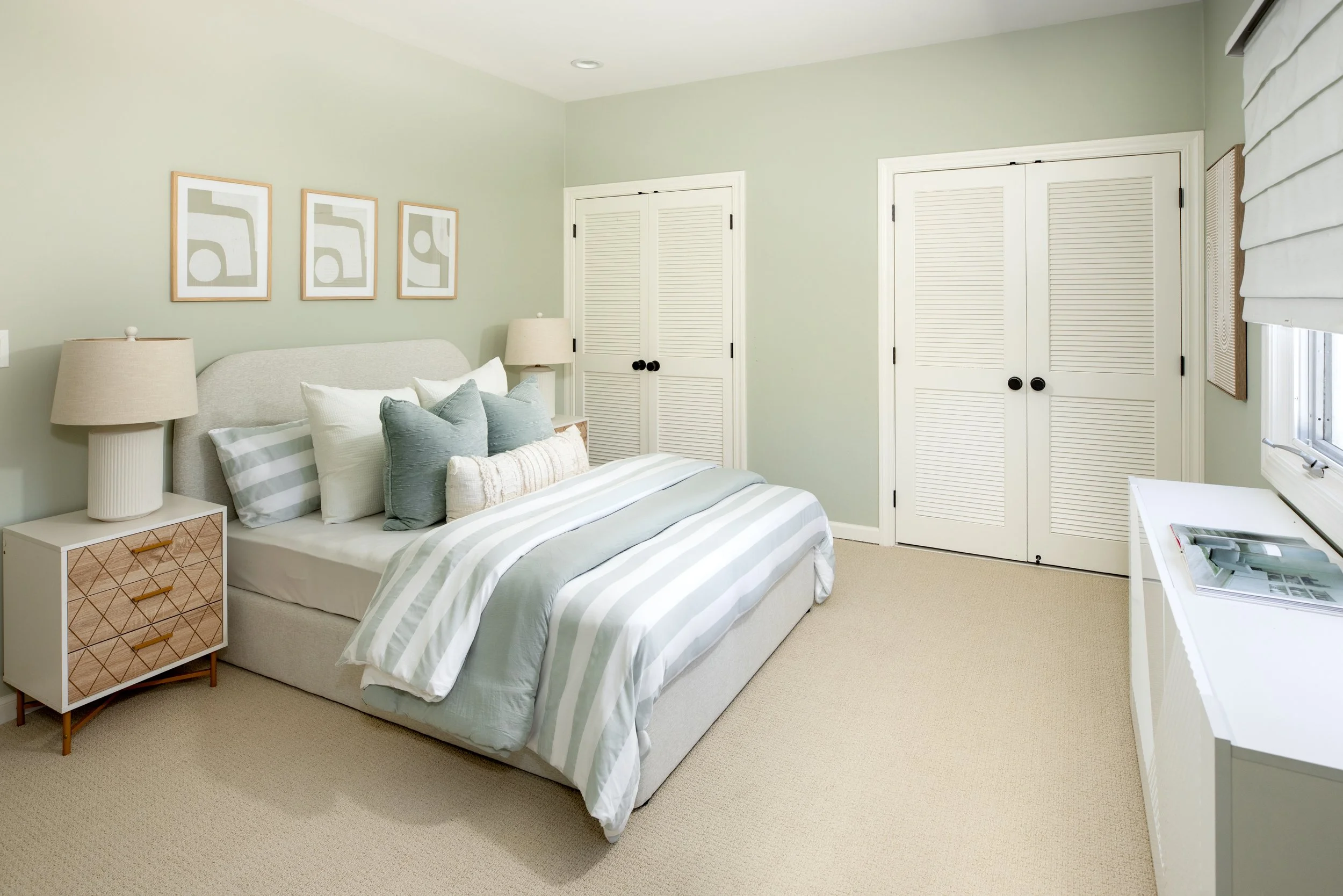 A bedroom with a bed, nightstand, and lamps. Light green walls, framed abstract art, closet with louvered doors, window with blinds, and beige carpet.
