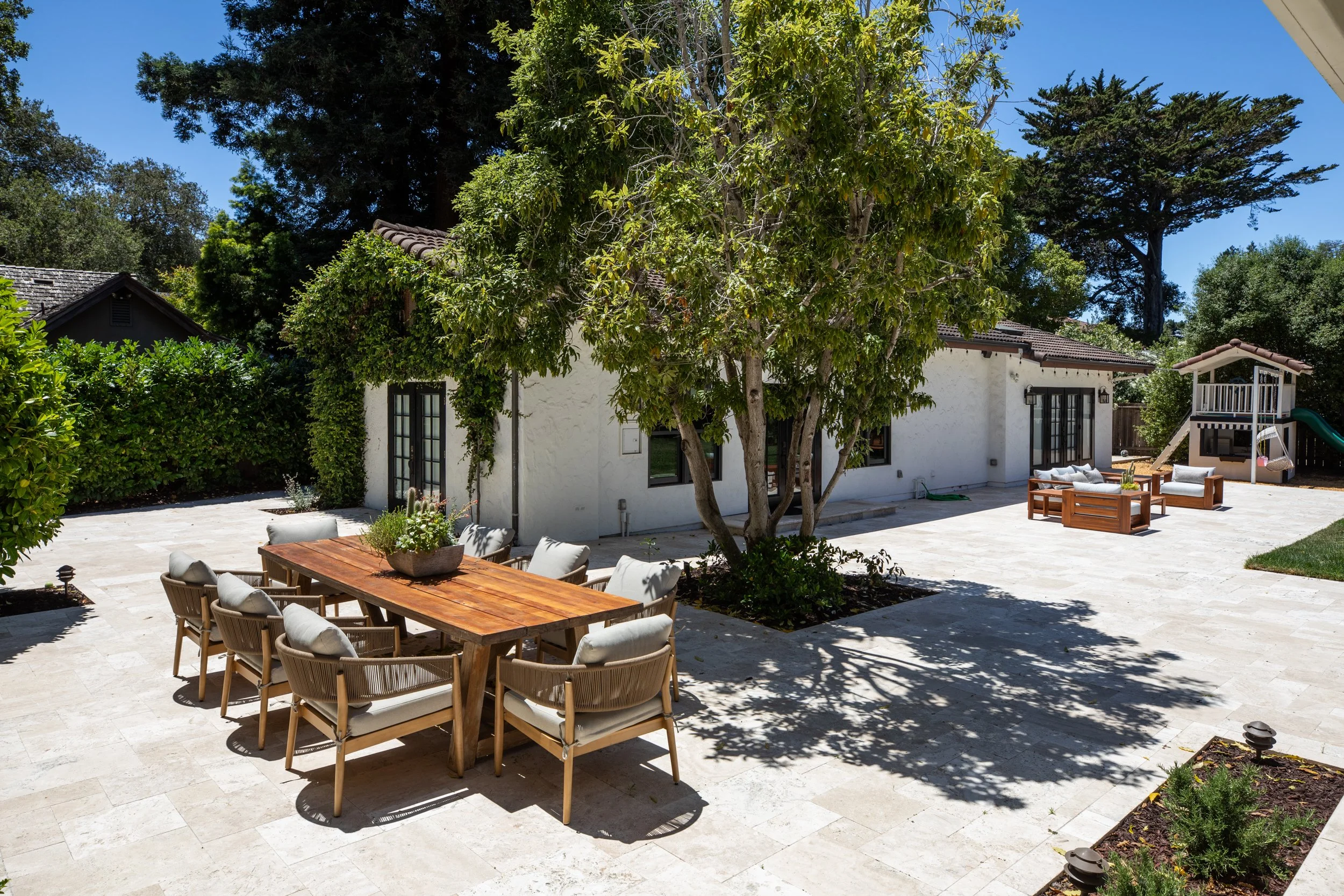 A backyard patio area with a white house, outdoor furniture including a dining table with chairs, and a playset with a slide, surrounded by trees and greenery under a clear blue sky.