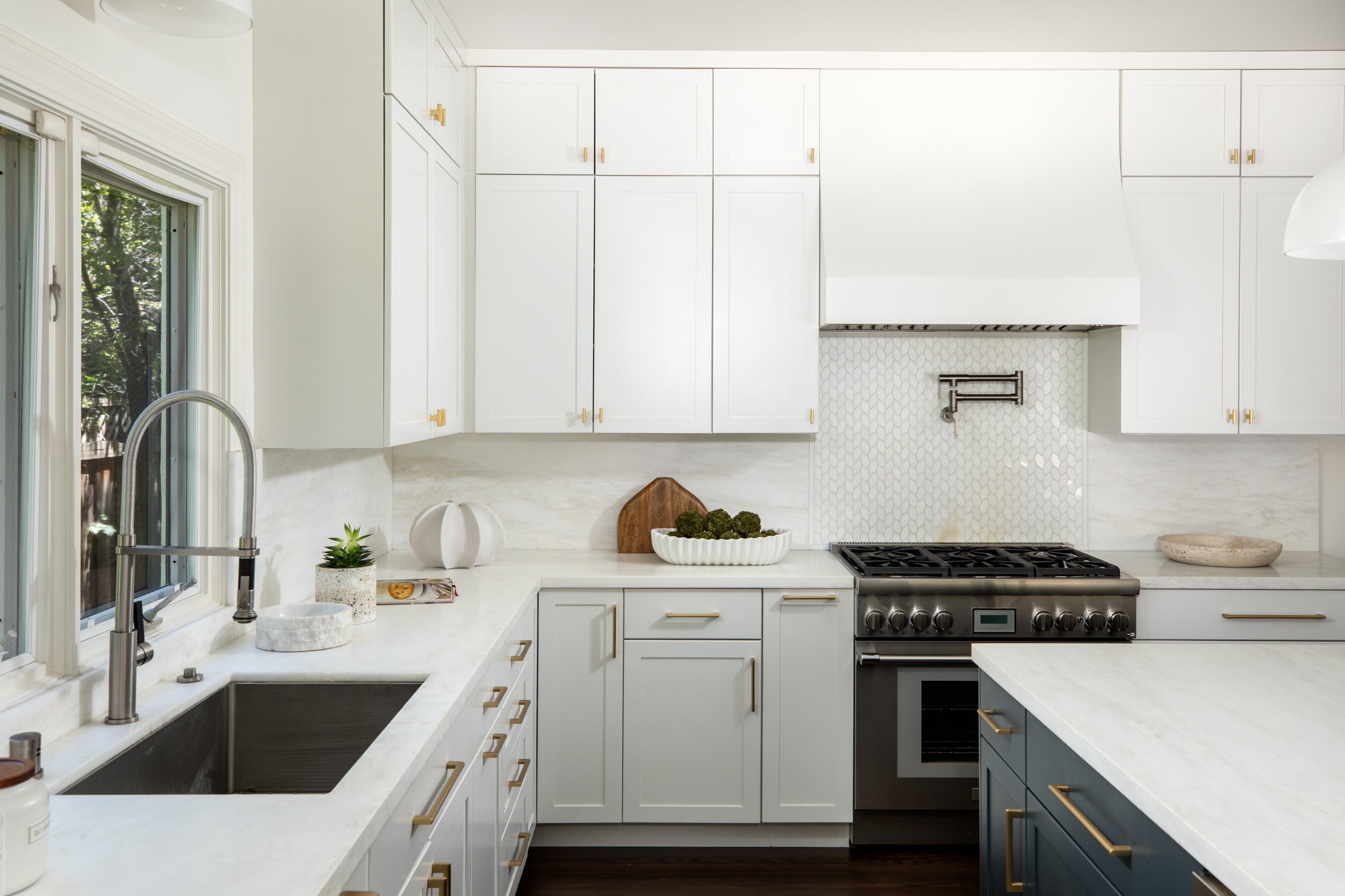 A modern kitchen with white cabinets, gold handles, a marble countertop, and a mosaic tile backsplash. There is a window with a view of trees, a double sink with a tall faucet, and a gas stove with an oven. Decorative items include a small potted pla