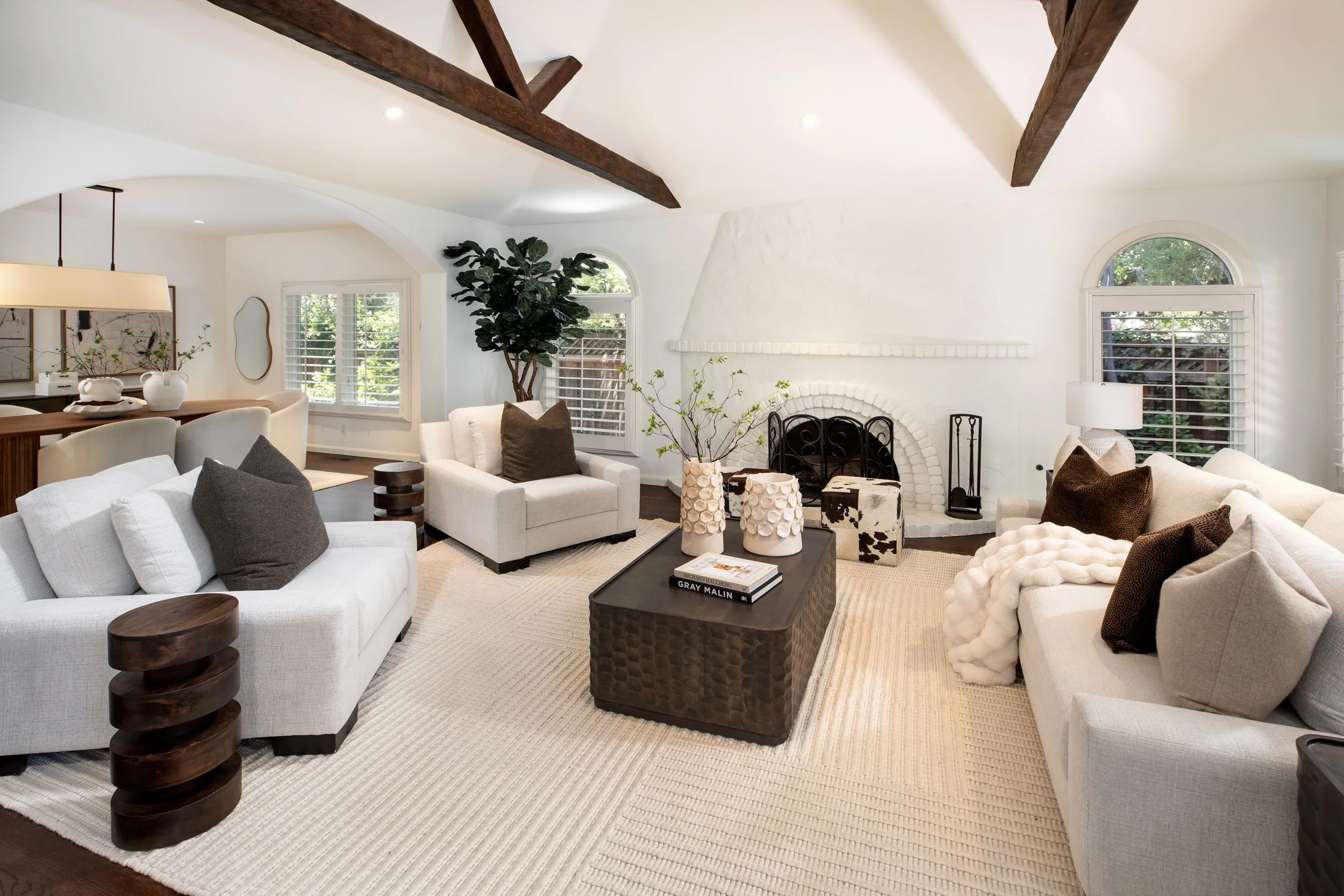 Living room with white sofas, dark wood side tables, a black coffee table, and beige areas rug. Large windows with white shutters, a white brick fireplace, indoor plants, and wall art.