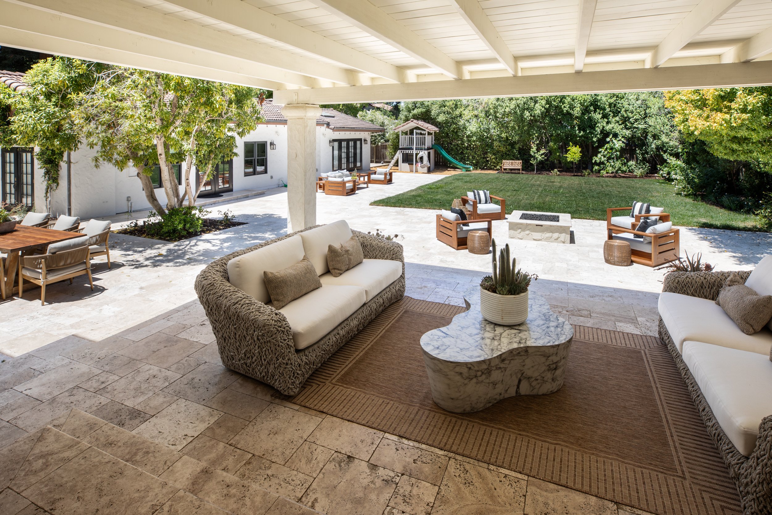 A spacious outdoor patio area with comfortable seating, a firepit, and a grassy yard with a play structure and trees.