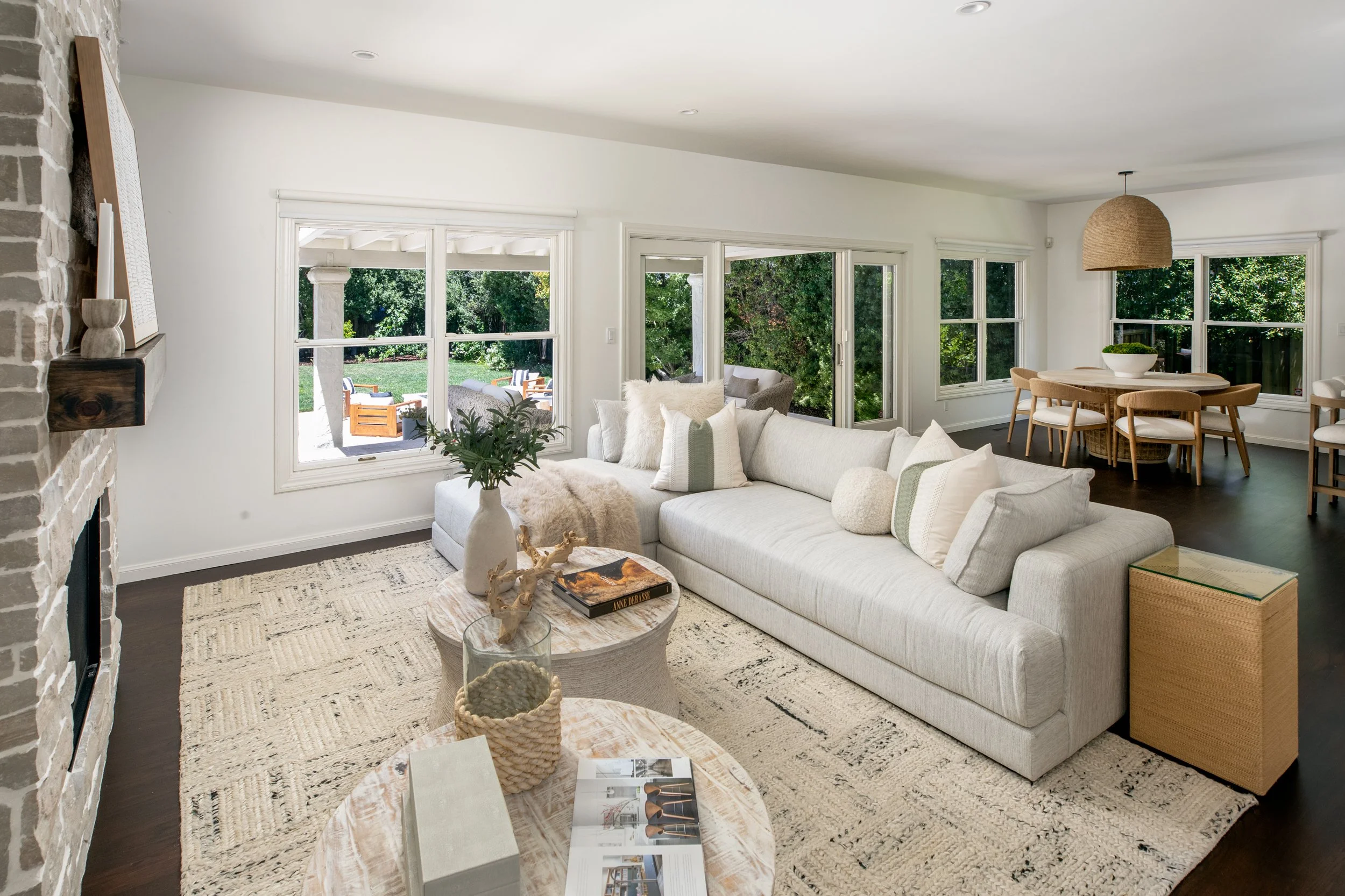 Living room with beige sectional sofa, round marble coffee table, textured white rug, and large windows with a view of a lush green backyard with outdoor furniture.