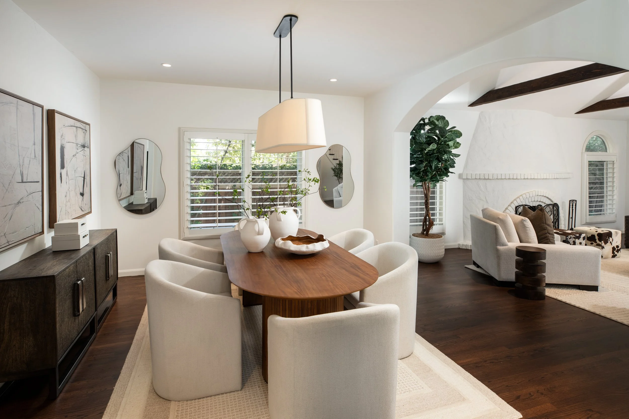 A living and dining room with white walls, dark hardwood floors, and large windows with plantation shutters. The dining area has a wooden table surrounded by white upholstered chairs, a hanging pendant light, and decorative vases with branches. The l