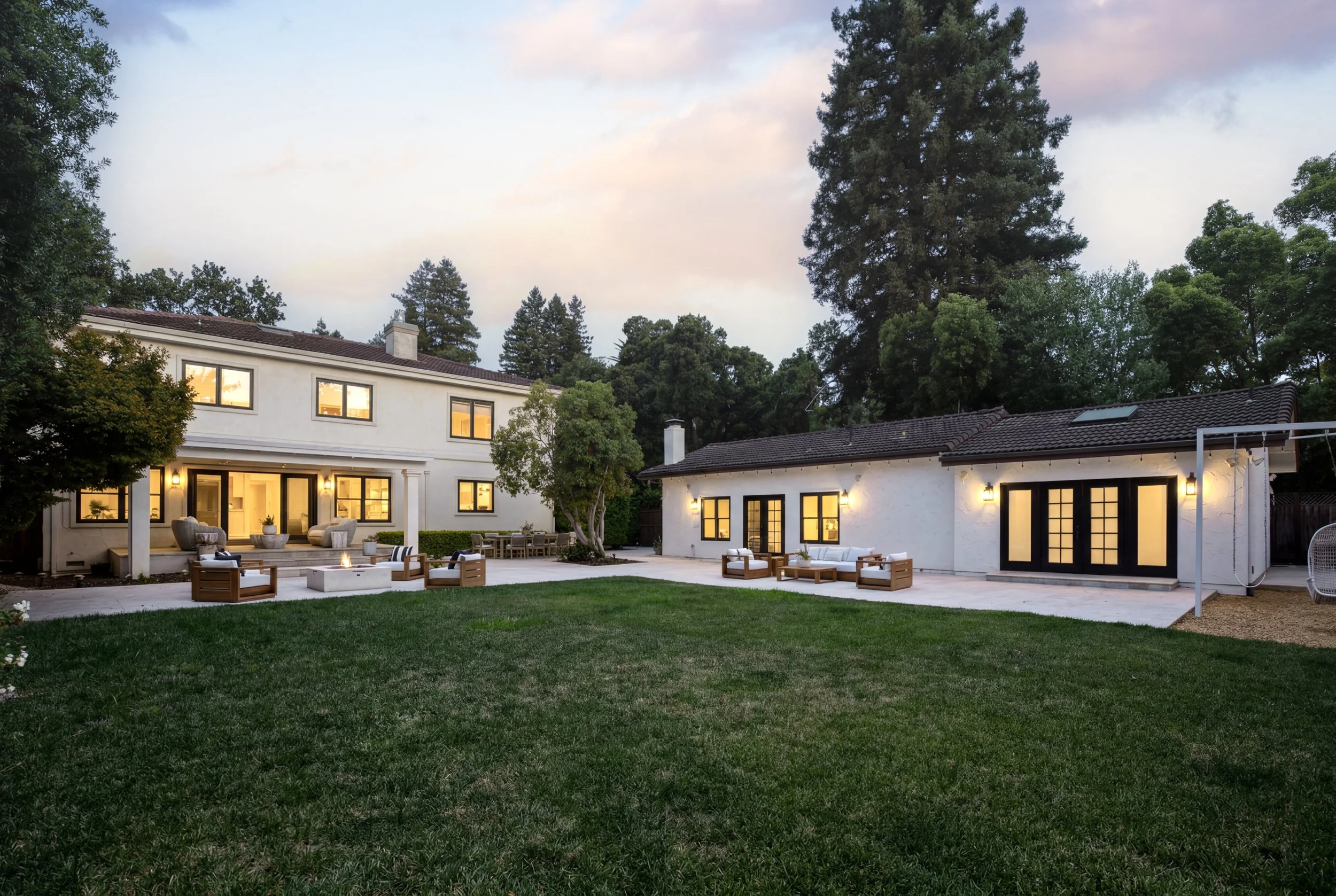 Backyard view of a modern white house at dusk with outdoor seating, fire pit, trees, and a large grassy lawn