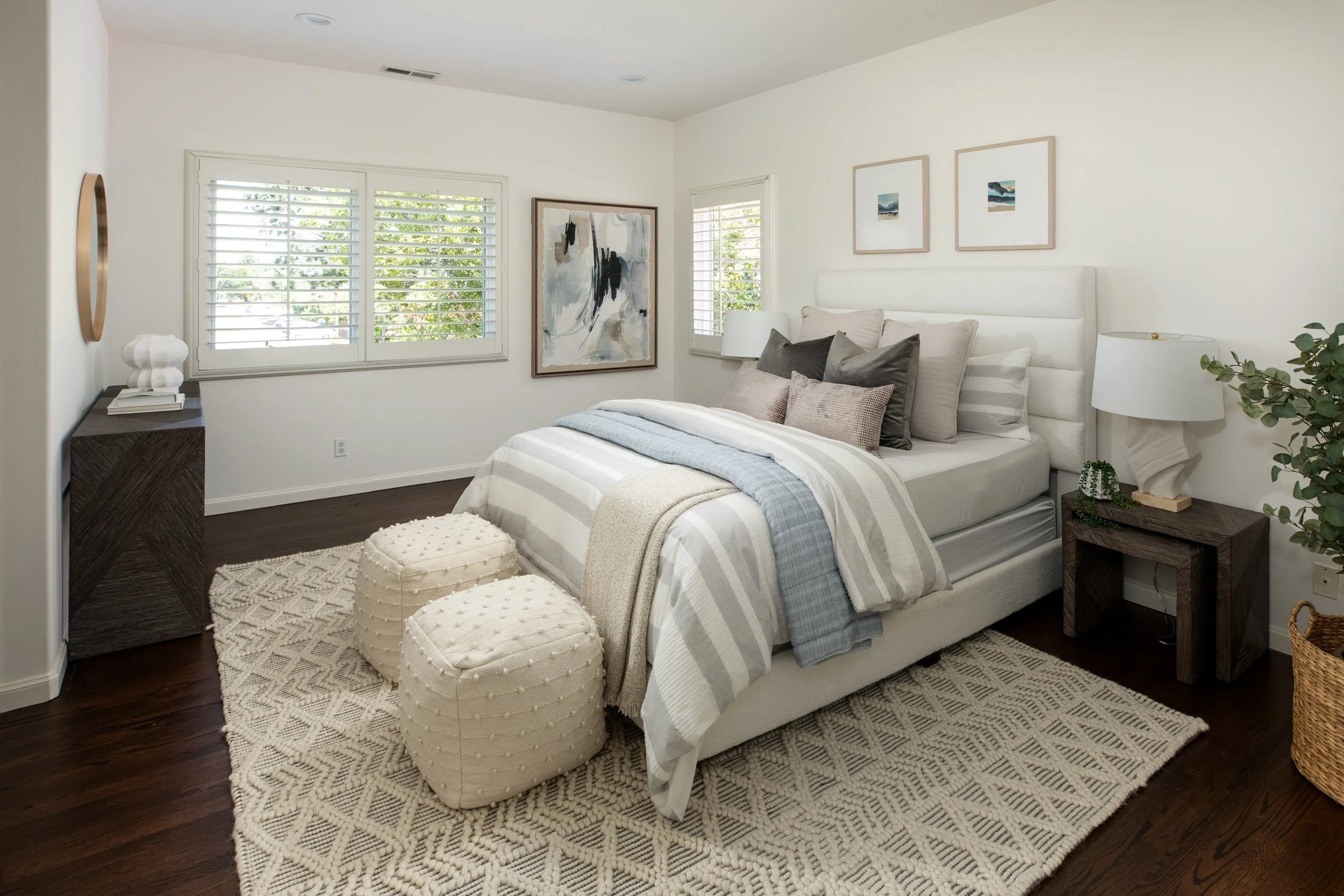 A bedroom with a white bed, multiple pillows, and a striped bedspread. There are two round, cream-colored ottomans at the foot of the bed, a dark wood nightstand with a white lamp and a small plant, and framed artwork on the walls. Two large windows 